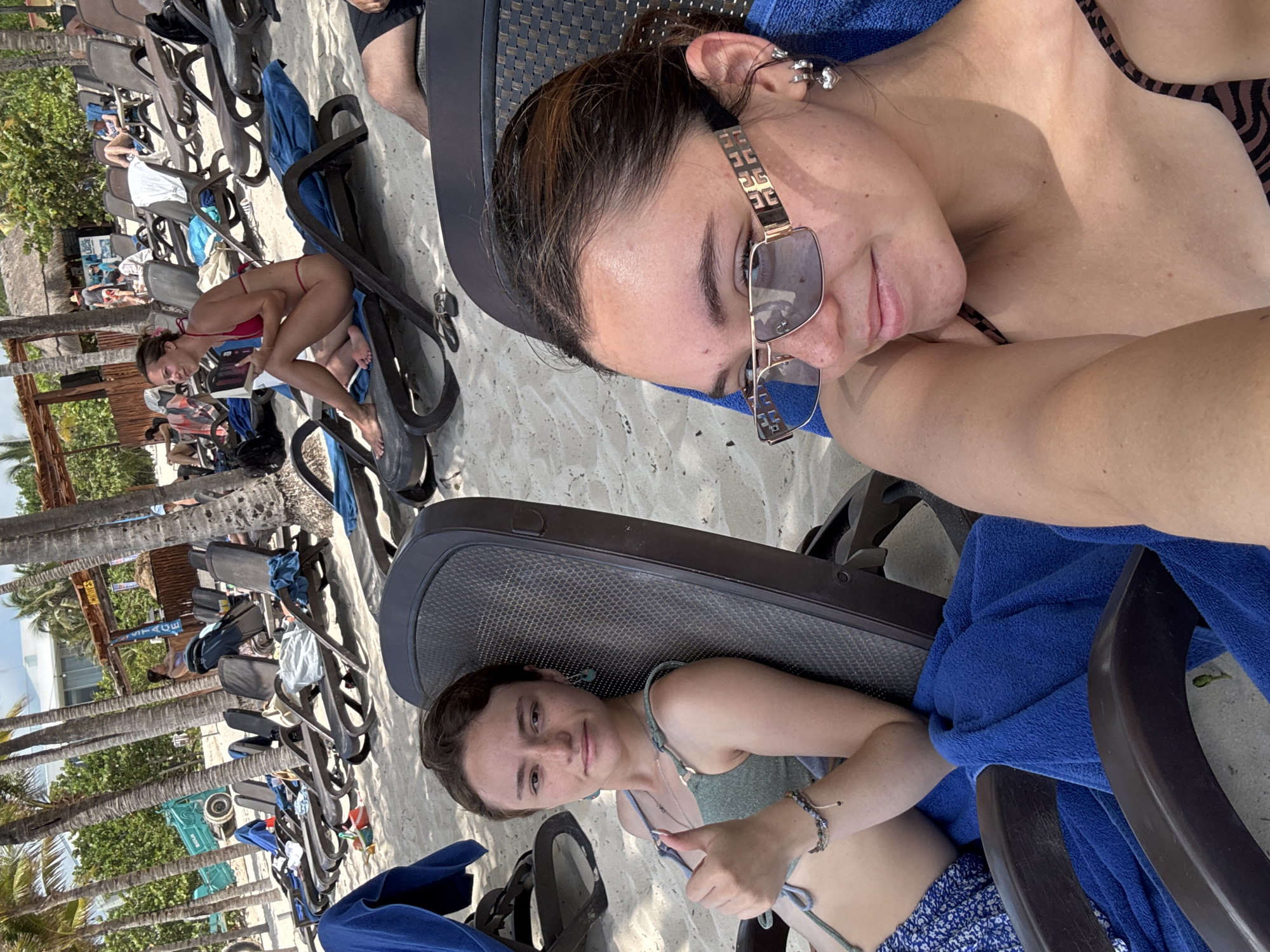 The image shows two women relaxing in beach chairs on a sandy beach with palm trees in the background. The woman in the foreground is wearing sunglasses and appears to be lying down. The second woman is seated and looking at the camera. Further away, a third woman in a red bikini is sitting in a beach chair, reading a book. Other beach chairs and people are visible in the background. Given the presence of palm trees, sandy beach, and beachside infrastructure, the location could be a resort or public beach in Playa del Carmen, Mexico.