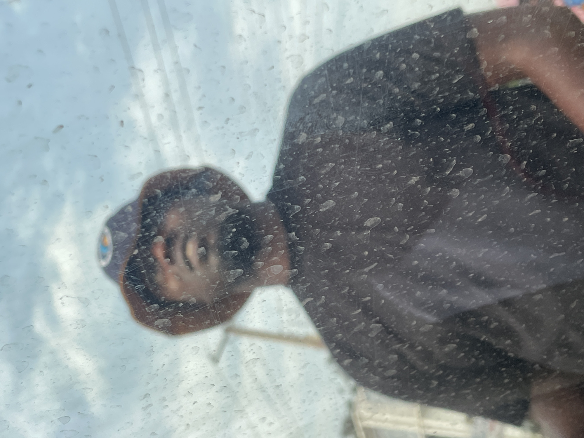 A person is reflected in a dirty glass surface. The person is wearing a brown hat and a dark-colored shirt. The reflection also shows a cloudy sky and a building with a white facade. The location is Jos, Nigeria.