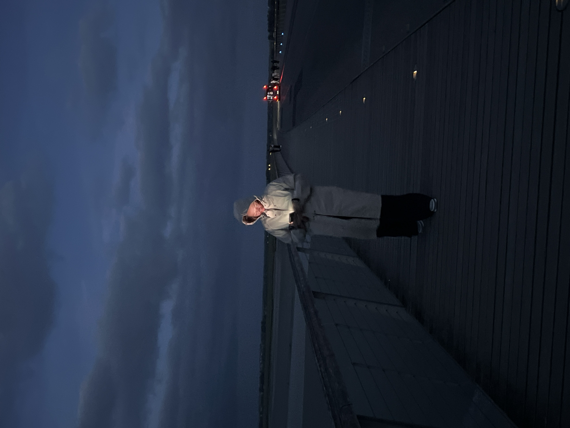The image captures a nighttime scene on the elevated causeway leading to Mont Saint-Michel, France. In the central foreground, a person wearing a light-colored hooded raincoat and dark trousers walks on a wooden-planked pathway, their face illuminated by the bright screen of a smartphone held in their hands. To the left, a dark railing borders the pathway, beyond which lies the expansive, indistinct tidal flats or bay of Mont Saint-Michel, meeting a low horizon of land under a dark, heavily clouded sky. The wooden pathway features small, embedded lights illuminating the ground. To the right of the person, a wider paved road or causeway extends into the distance, also fitted with ground-level lights. A red vehicle, possibly a shuttle bus, with illuminated rear lights and other white lights, is visible in the mid-ground on this road, accompanied by several small, unidentifiable human figures. The overall environment is dark, with ambient light provided by the sky's faint glow, the phone screen, embedded walkway lights, and vehicle lights.