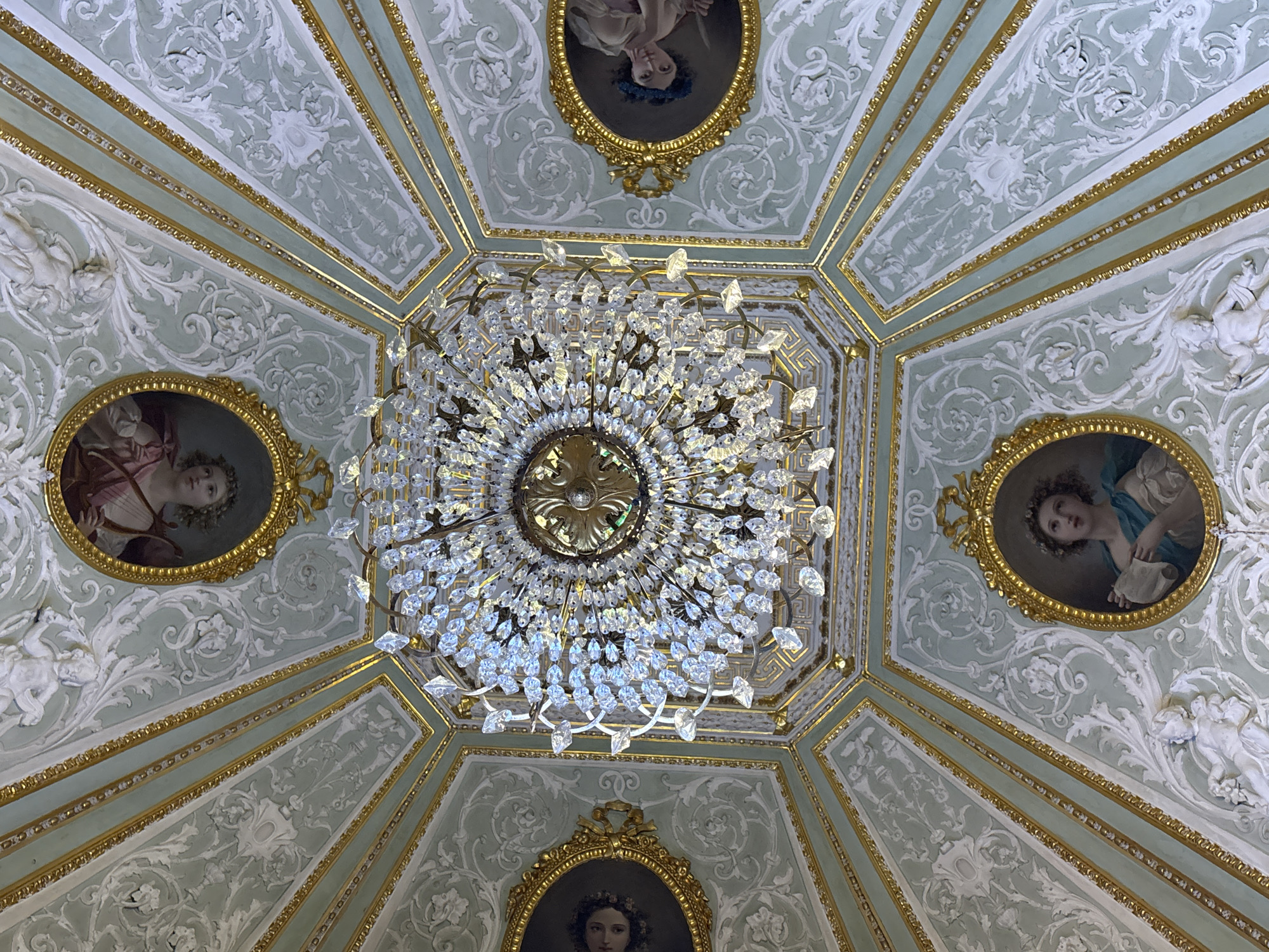 This image captures an ornate, octagonal ceiling in Saint Petersburg, Russia, likely within a historical palace or museum. Dominating the center is a large, multi-tiered crystal chandelier, its facets catching and reflecting light. The ceiling itself is decorated with a pale mint green background and intricate white stucco moldings featuring floral and scrollwork motifs. Several oval portraits in gilded frames are set into the ceiling panels, each depicting a young woman. The portraits exhibit classical styling with soft lighting and detailed rendering of facial features and clothing. The architecture suggests a lavish, pre-revolutionary Russian interior.