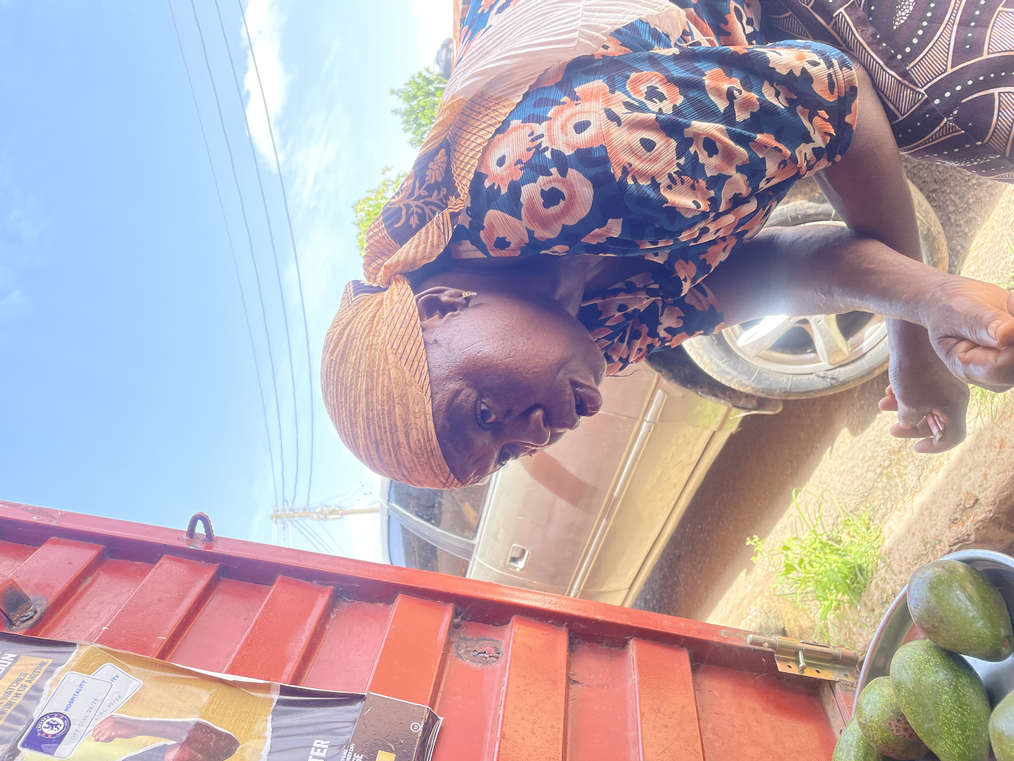Here's a detailed description of the image:
**People:**
* An African woman with a brown head wrap, and floral print dress.
**Subjects:**
* A container with a red exterior.
* A silver car.
* A bowl of avocados.
* Posters attached to the side of the container.
**Objects:**
* Avocados in a bowl
* Posters with text and images
* Silver car
* Red container
**Actions:**
* The woman is looking down at the avocados and the car is in the background.
**Interactions:**
* The woman appears to be selling avocados from the container.
**Environment:**
* The location appears to be outdoors, possibly a roadside.
* The sky is visible in the background with power lines.
* There appears to be a concrete block under the wheel of the vehicle
*The context of this image may be Jos, Nigeria.