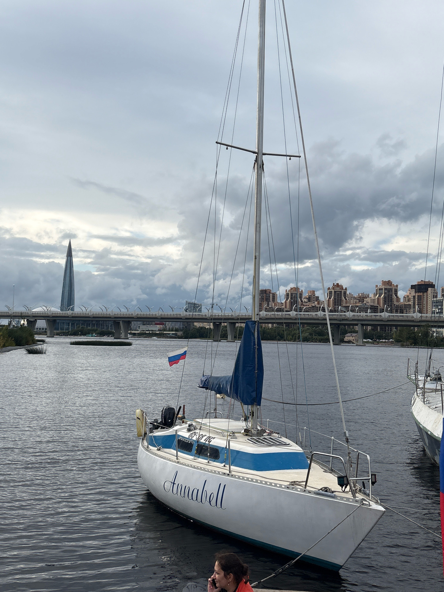 The image depicts a cloudy day scene on a body of water, likely a river or bay, in Saint Petersburg, Russia. In the foreground, a white sailboat with blue trim is moored. The name "Annabell" is written in blue script on its hull. A small Russian flag is visible on the boat. In the background, a modern bridge with distinctive arch supports spans the water, with a unique, tall, spire-like skyscraper visible to the left of the bridge. Several apartment buildings are visible beyond the bridge. The overall atmosphere is overcast and somewhat muted, characteristic of Saint Petersburg's climate. A person is partially visible on the dock in the bottom right corner, appearing to be on a phone.
