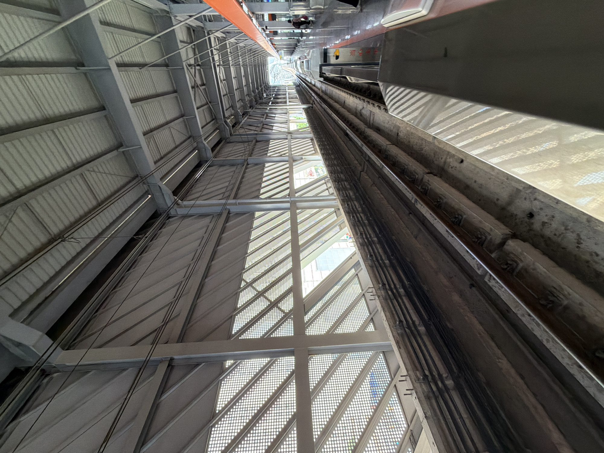 The image depicts a view from a train platform looking down the train tracks in Tiaodeng, China. The platform is covered by a modern, angular roof structure supported by metal beams. The walls of the structure are made of a material with a diamond-shaped grid pattern, allowing light to filter through. Train tracks run along the platform and extend into the distance. On the right side of the platform, there is a person sitting down, and further down the platform, another person is standing. The overall environment is a covered train station, characterized by industrial design and construction.