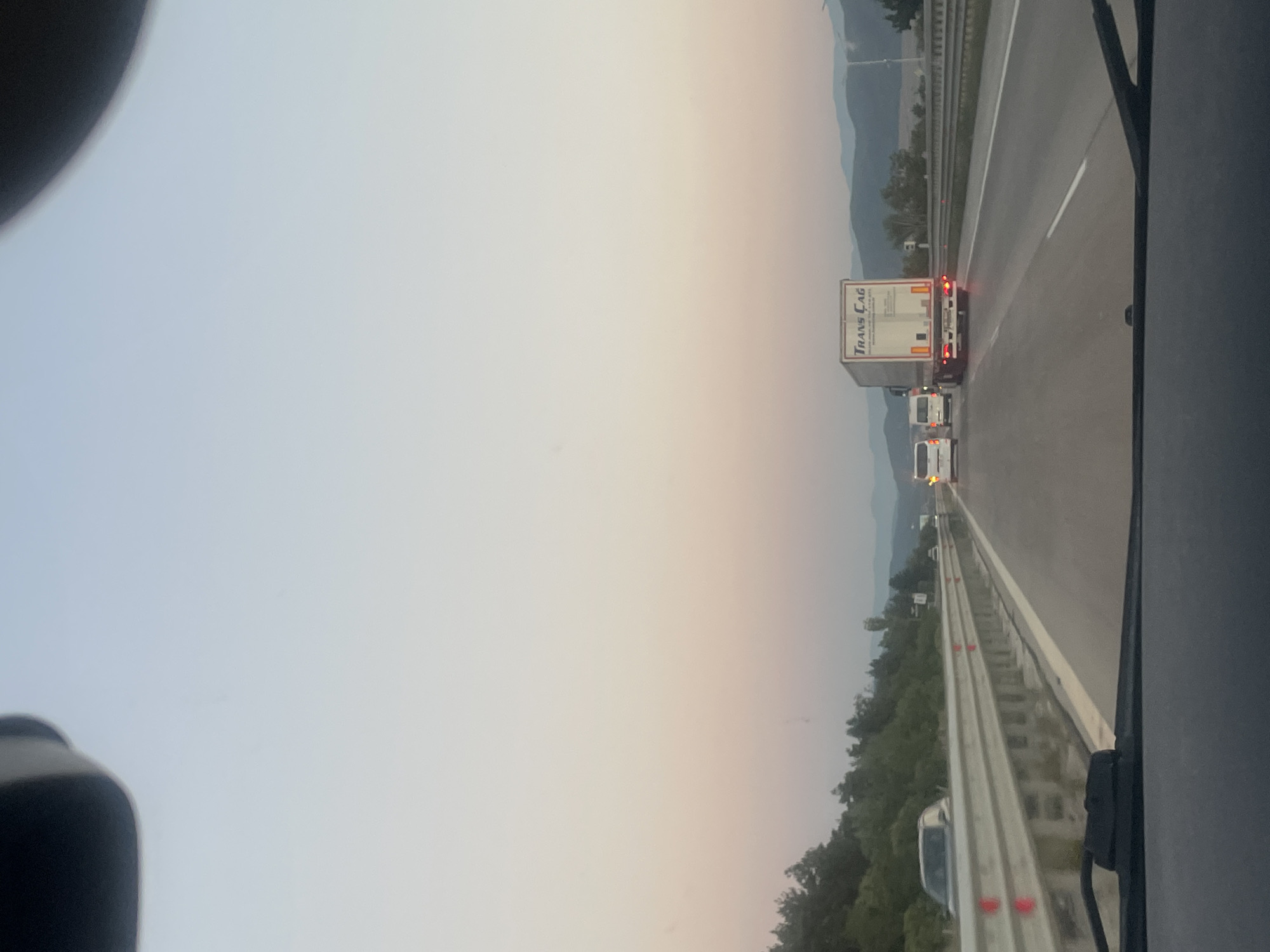 The image shows a view from a vehicle on a highway, likely the A1 highway near Ihtiman, Bulgaria, at dusk. The sky is a gradient of soft pinks, oranges, and blues. In the foreground, the dashboard and windshield wipers of the vehicle are visible. The highway has multiple lanes separated by guardrails and white lines. Several vehicles are on the road: a white truck with "TRANS ÇAG" written on its back, two white vans, and a dark-colored car in the left lane. Trees and hills form the landscape on the sides of the highway, with mountains visible in the distance. The overall atmosphere is calm and serene, characteristic of a sunset drive.