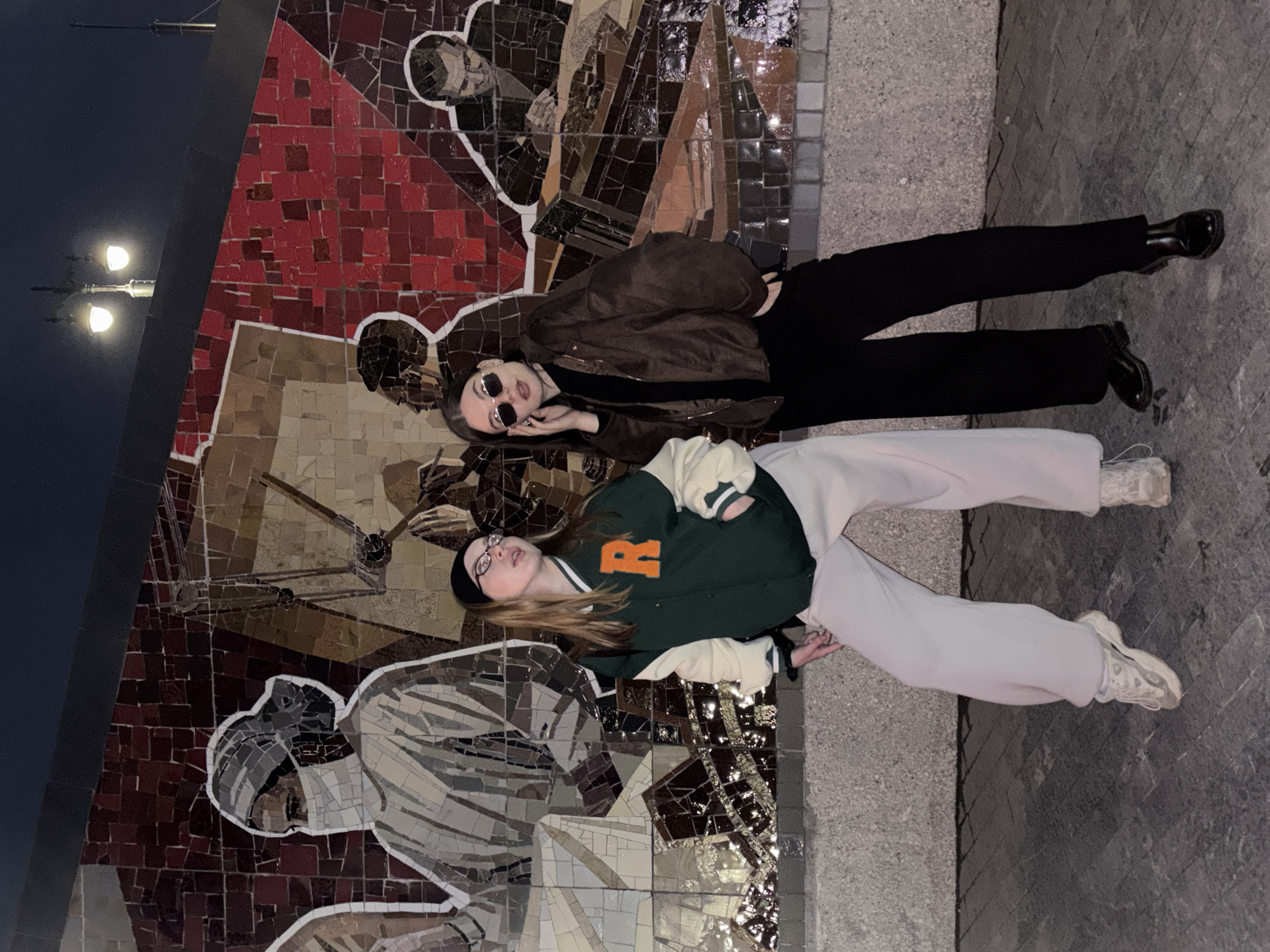 Two young women are reclining on a mosaic mural. The mural depicts figures in a Soviet Realism style. The woman on the bottom is wearing a dark beanie, glasses, a dark green letterman-style jacket with a large orange "R," and light-colored sweatpants with white sneakers. The woman above her is wearing small round sunglasses, a brown jacket, a black shirt, black pants, and black boots. The location is Perm, Russia; the mural is a prominent feature of the city, and it is on a platform of patterned concrete. The lamppost is in the background.