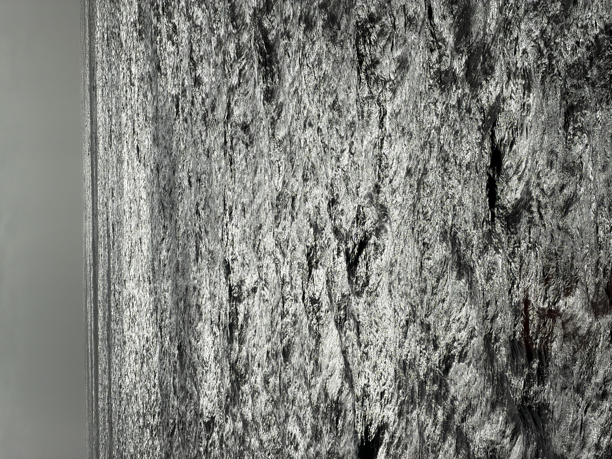 The image depicts a large, dark, textured rock face or cliff. The rock appears rough and uneven, with various ridges, crevices, and shadowed areas that create a complex surface pattern. The left side of the image reveals a lighter, overcast sky, indicating the presence of a horizon line. The absence of any discernible objects or subjects within the image suggests a natural, unpopulated environment.