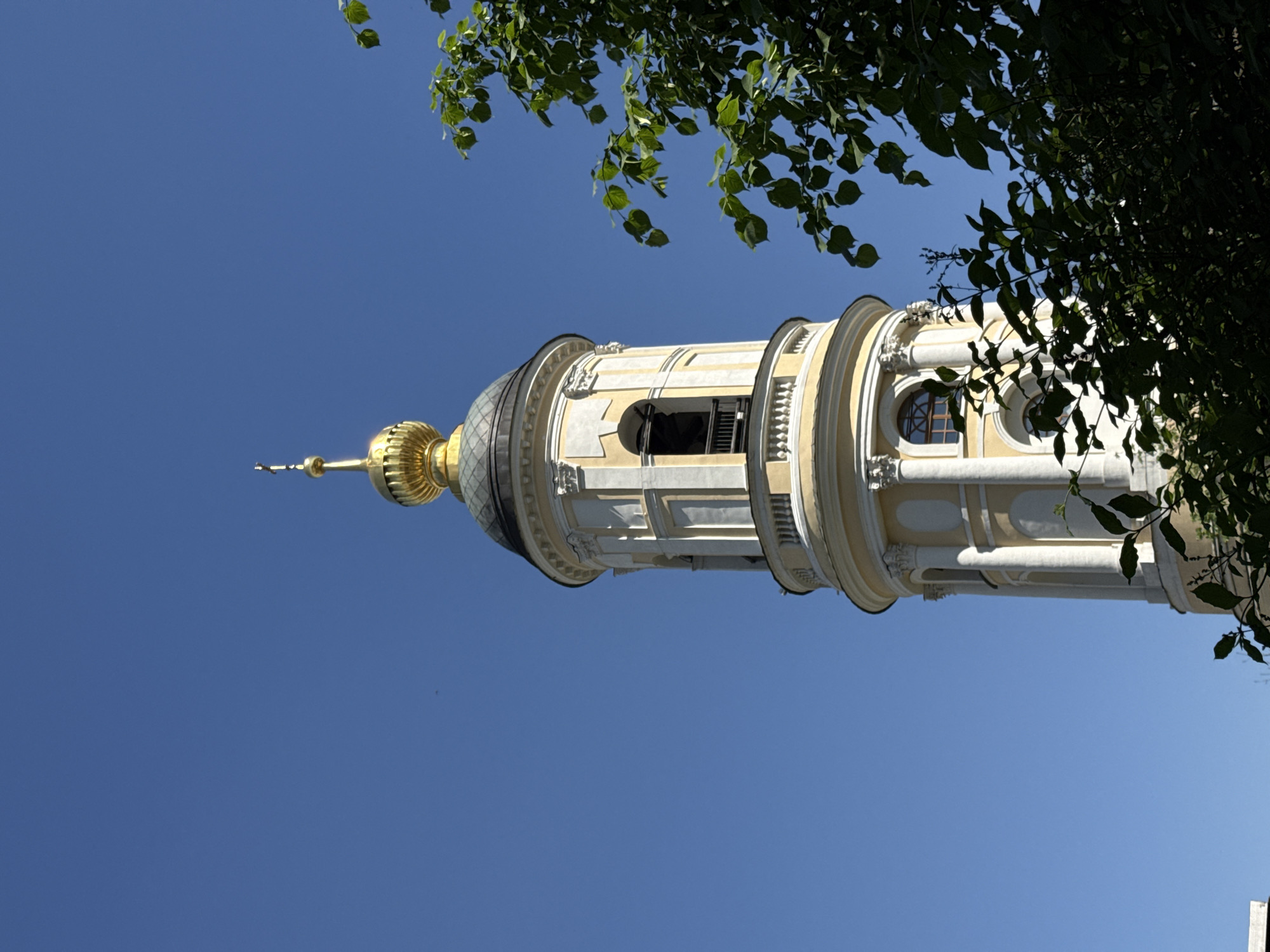 Here is a description of the image:
The image shows the upper portion of a tall, ornate bell tower against a clear blue sky. The tower is predominantly white with golden accents at its dome and spire. Its architecture features multiple tiers separated by decorative moldings and rows of arched windows, some of which have black grates. The right side of the image is partially obscured by the dense foliage of a tree, creating a frame of green leaves around the tower. Some tree branches extend from the top left, enhancing this framing effect. The tower's vertical orientation dominates the composition, with the sky serving as a uniform, contrasting backdrop.