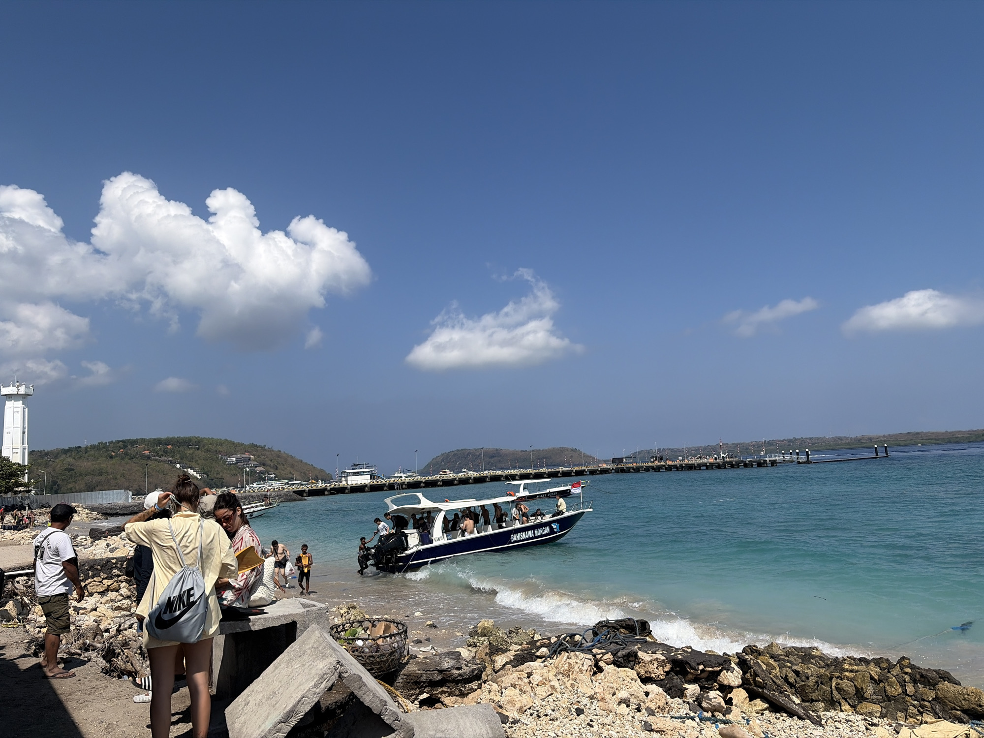 In Nusa Penida, Indonesia, a bright, sunny day at a harbor features a bustling scene of activity. A blue and white speedboat, labeled "BAHISHAWA MORGAN," cruises in the turquoise water near the shore, carrying passengers. People are seen disembarking from or preparing to board the boat, with some wading into the shallow water. The shoreline is a mix of sand and rocky debris, with concrete structures scattered along the beach. In the background, a long pier extends into the sea, with buildings and lush green hills visible on the distant island. A white lighthouse stands prominently on the left side of the image, adding to the coastal landscape. Several individuals are gathered on the land, some appearing to be tourists with bags and casual attire, while others, possibly locals or workers, are also present. The sky is a clear, vibrant blue, dotted with fluffy white clouds.
