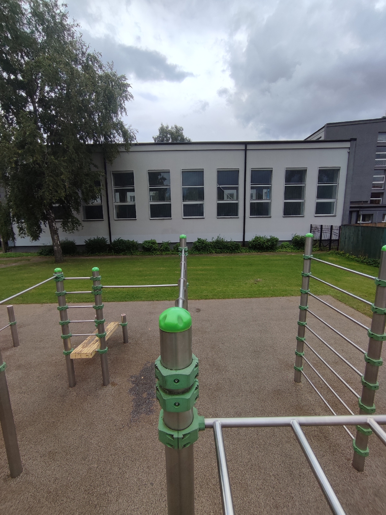 The image captures an outdoor exercise area with metal bars and a wooden bench, set against a backdrop of a white building with multiple windows. The ground is covered in a textured, light brown material, and a patch of green grass is visible behind the exercise equipment. A tall tree with green foliage stands to the left of the building. The sky is overcast with grey clouds. The exercise equipment consists of vertical silver poles with green connectors and horizontal silver bars. One piece of equipment features a wooden bench supported by silver poles. The image appears to be taken during the daytime under a cloudy sky. This scene is located in Rīga, Latvia.