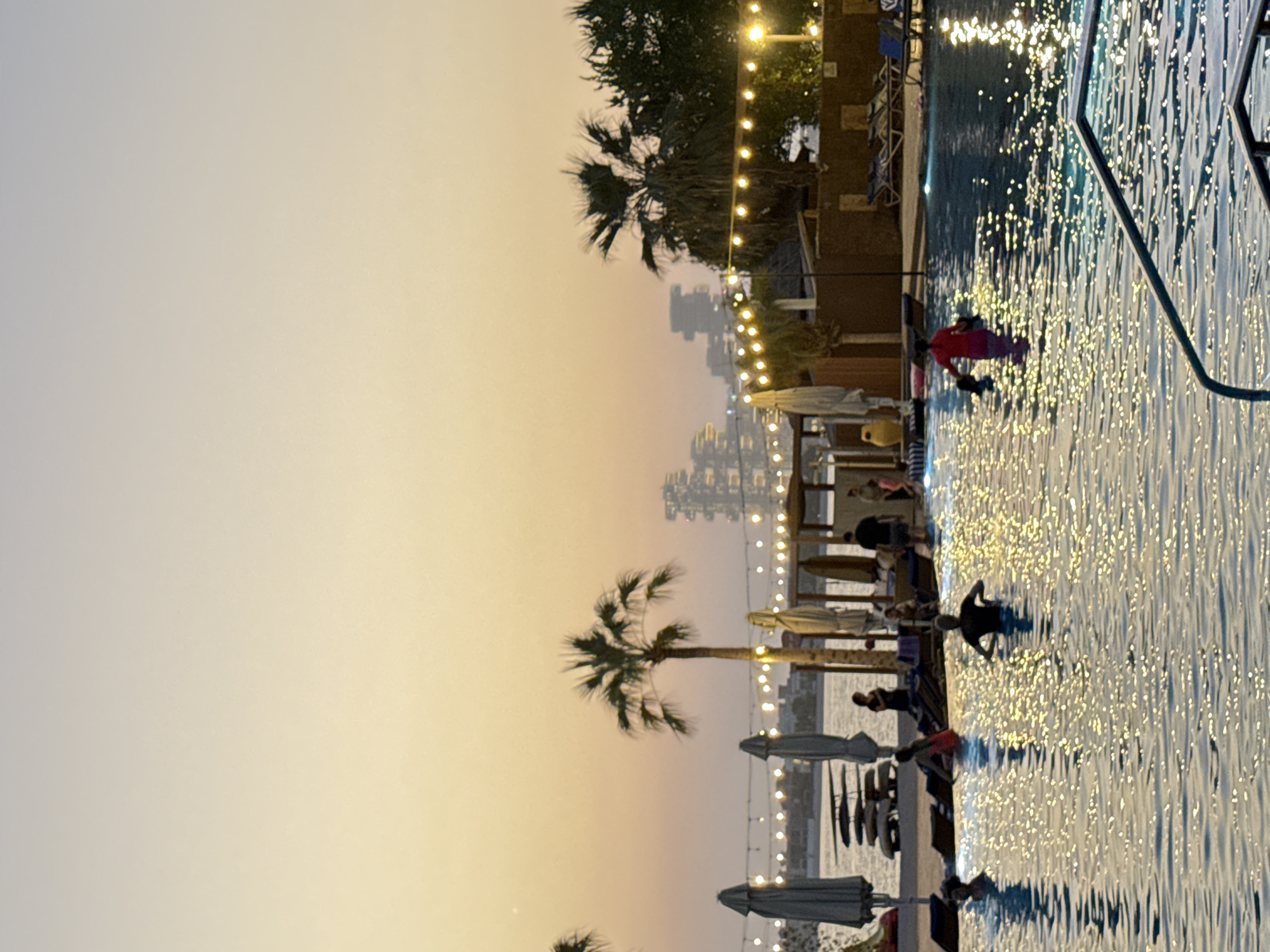 The image depicts a twilight scene at a resort-style swimming pool and beach area in Dubai, United Arab Emirates. The sky exhibits a gradient of soft orange and yellow hues, indicative of sunset or sunrise. In the foreground, a large swimming pool is occupied by several individuals, some partially submerged. The water's surface reflects the ambient light, creating a shimmering effect. Beyond the pool, lounge chairs and umbrellas are arranged on a sandy area, suggesting a beach setting. Palm trees are visible, along with string lights that add to the ambiance. In the background, a hazy silhouette of buildings suggests a city skyline, characteristic of Dubai's urban landscape. The overall impression is one of a relaxed evening at a tropical resort.