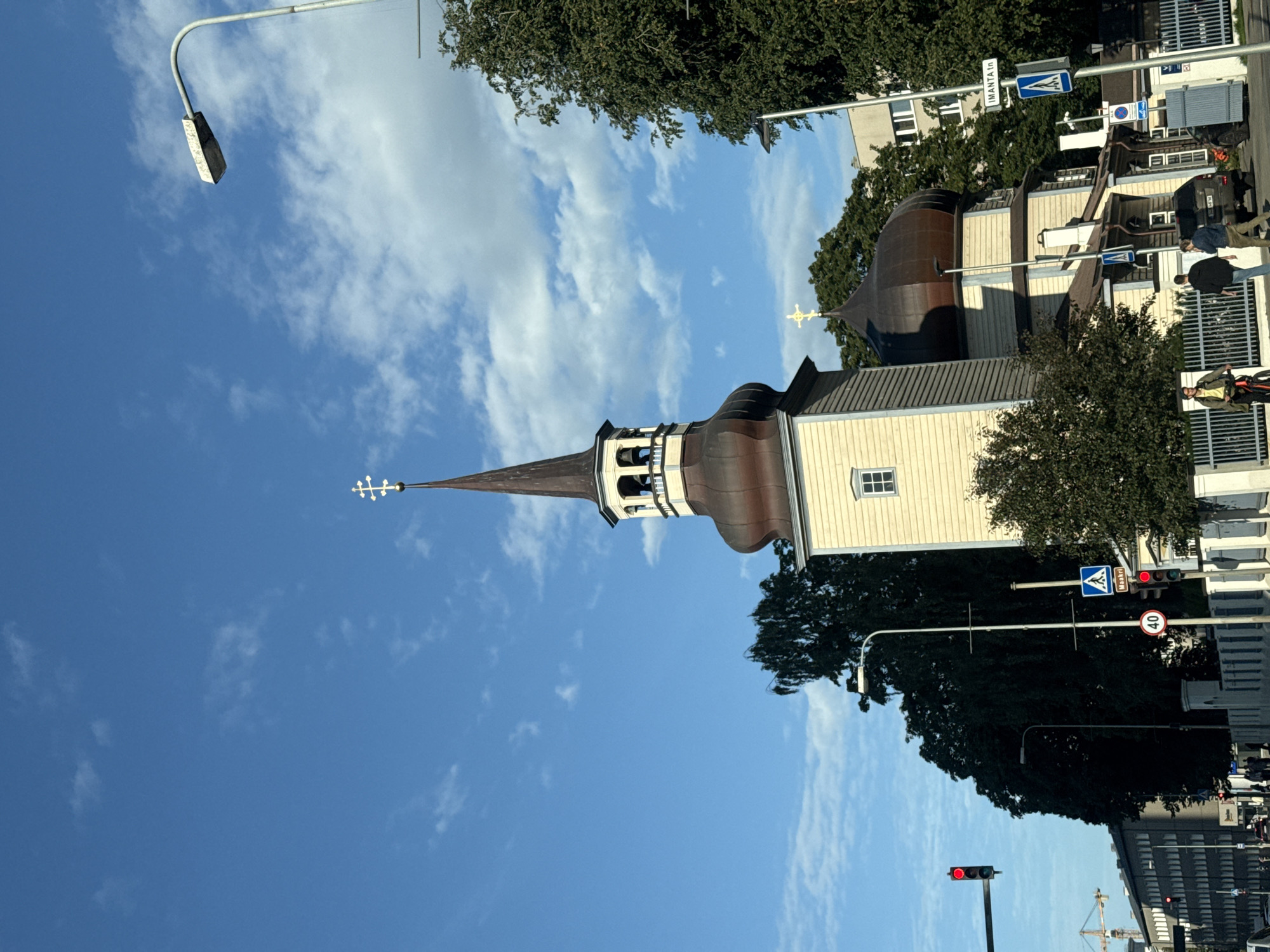 In Tallinn, Estonia, a light-colored wooden church with a tall, dark steeple topped with a golden cross stands against a bright blue sky with scattered clouds. The church has multiple domed roofs and wooden siding. In the foreground, a paved road is visible with pedestrian crossings marked by blue and white signs. Traffic lights are present, one showing a red signal. Several trees, both deciduous and evergreen, surround the church and the roadside. A street lamp with a rectangular fixture is positioned on the right side of the image. In the background, a multi-story building and a construction crane suggest an urban environment. Several people are walking on the sidewalk and near the road. Two individuals are seen walking away from the camera, and another person is on a bicycle. Vehicles are present on the road. The overall scene depicts a street view in Tallinn on a clear day.