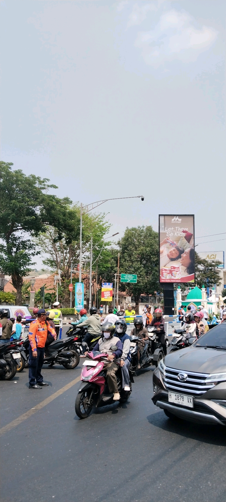 The image depicts a busy intersection in Semarang, Indonesia, with a significant amount of motorcycle traffic and a few cars. The scene is characterized by a traffic jam, with many people on motorcycles, some wearing helmets and masks. Traffic police officers in orange and yellow vests are visible, directing the flow of vehicles.
In the background, there are several billboards, including one that says "Let Them Be Kids." There are also trees, streetlights, and buildings, suggesting an urban environment. The sky is overcast, indicating a cloudy day. The overall impression is one of a typical day in a bustling Indonesian city, with the added element of traffic control possibly due to an event or just the usual congestion.