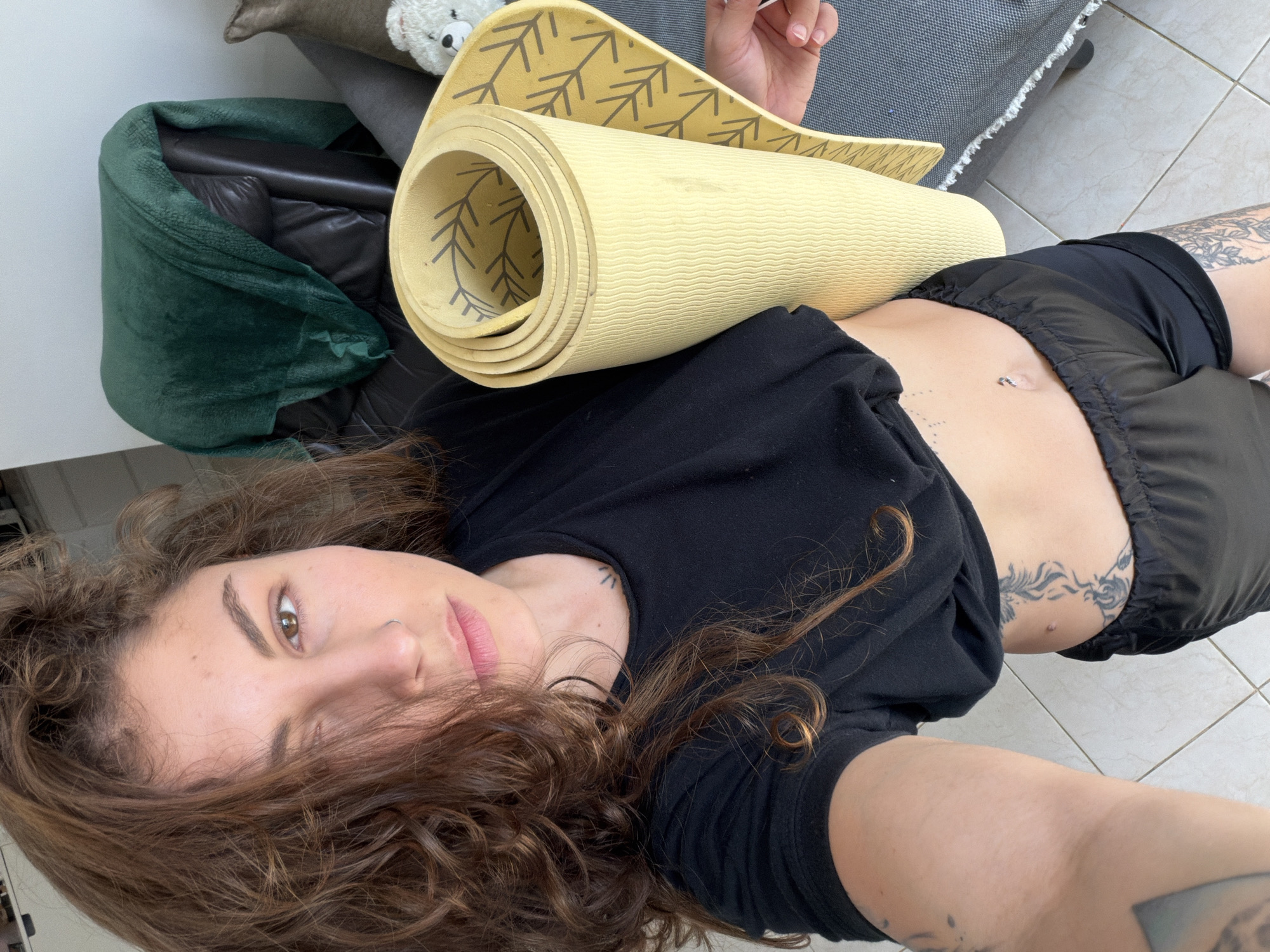 The image is a high-angle selfie of a young woman lying supine on a light-colored tiled floor in an indoor setting, likely Kiryat Bialik, Israel. She has long, curly brown hair framing her face, brown eyes, and a neutral expression, looking directly at the camera. She wears a black t-shirt and black athletic shorts.
Her visible features include a nose ring, a belly button piercing, and multiple tattoos: a prominent patterned design along her right rib cage/torso, and smaller, less distinct tattoos on her arms and shoulder.
A yellow yoga mat, partially unrolled and featuring a repetitive dark grey leaf or tree branch pattern, rests across her midsection. Her right hand is visible in the upper right corner, holding the mat.
The background reveals parts of a furnished room. To the left, a dark green, soft blanket is draped over a black armchair or sofa. Further back and to the right, a grey upholstered surface, possibly a couch or a large pet bed, is visible, with a small white stuffed teddy bear resting on it. The lighting suggests it is daytime. No discernible text is visible within the frame. The scene conveys a moment of rest or preparation related to physical activity.