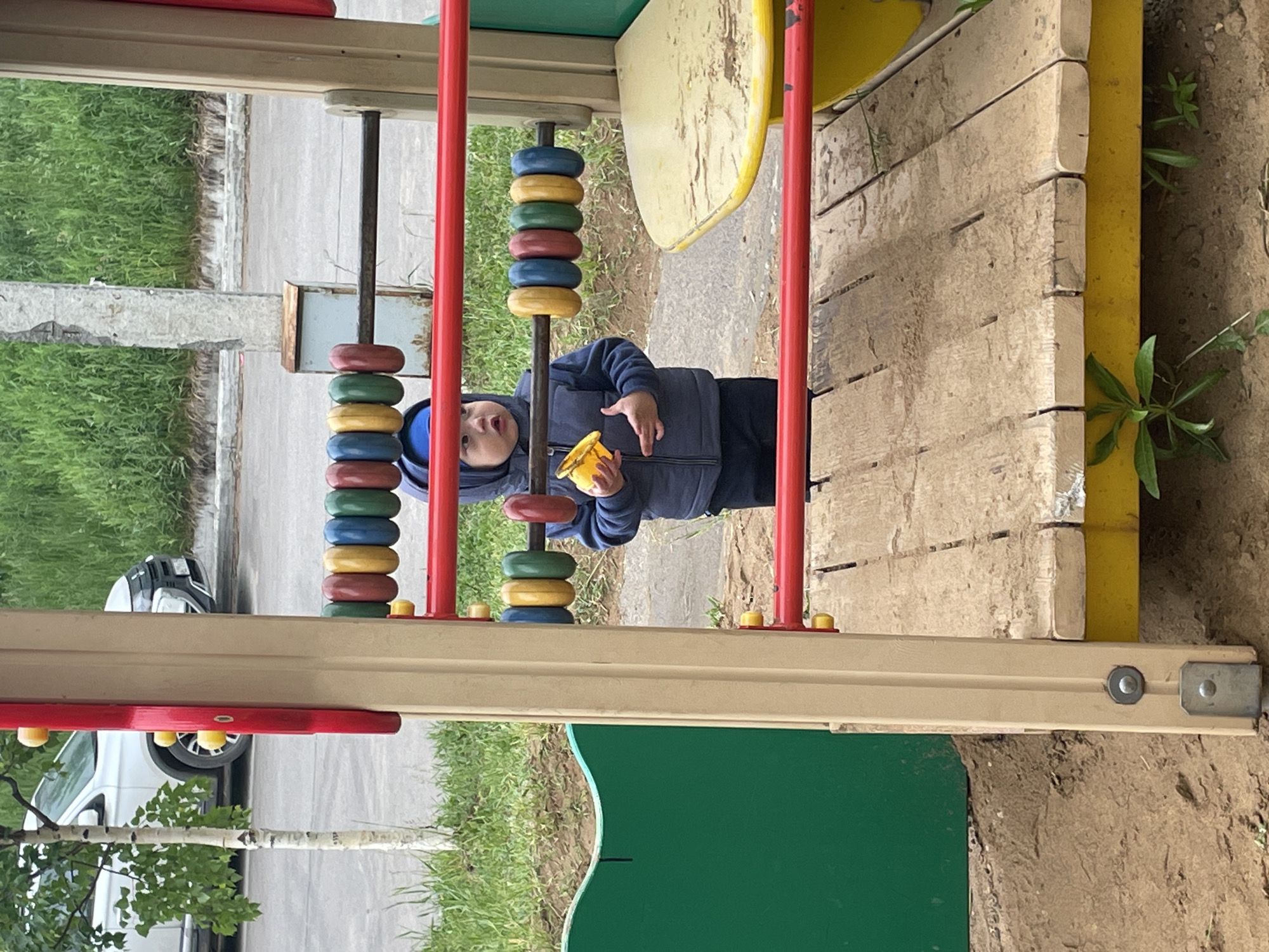 A young child is captured in an outdoor playground setting in Perm, Russia. The image is oriented with the playground equipment appearing vertical.
The central subject is a young child, likely a toddler, dressed in a blue hooded jacket or vest over a blue long-sleeved shirt and dark pants. The child is looking directly at the camera with a curious expression, holding a small yellow object, possibly a toy or snack, in their right hand.
The child is positioned behind a playground structure that includes an abacus-like feature with multiple colorful rings (red, green, yellow, blue, and brown) stacked on vertical poles. This feature is part of a larger, well-used playground made of wood and brightly colored plastic or painted metal elements, including red vertical supports, a yellow surface, and a green wavy panel.
In the background, beyond the playground, there is a paved surface, likely a sidewalk or road, bordered by green grass. A white car is partially visible on the left, parked on the paved area, with a tree featuring light-colored bark and green leaves in front of it. The ground immediately surrounding the playground structure appears to be dirt.
The scene is lit by natural daylight, suggesting it is daytime, possibly under an overcast sky as no harsh shadows are evident. There is no visible text in the image.