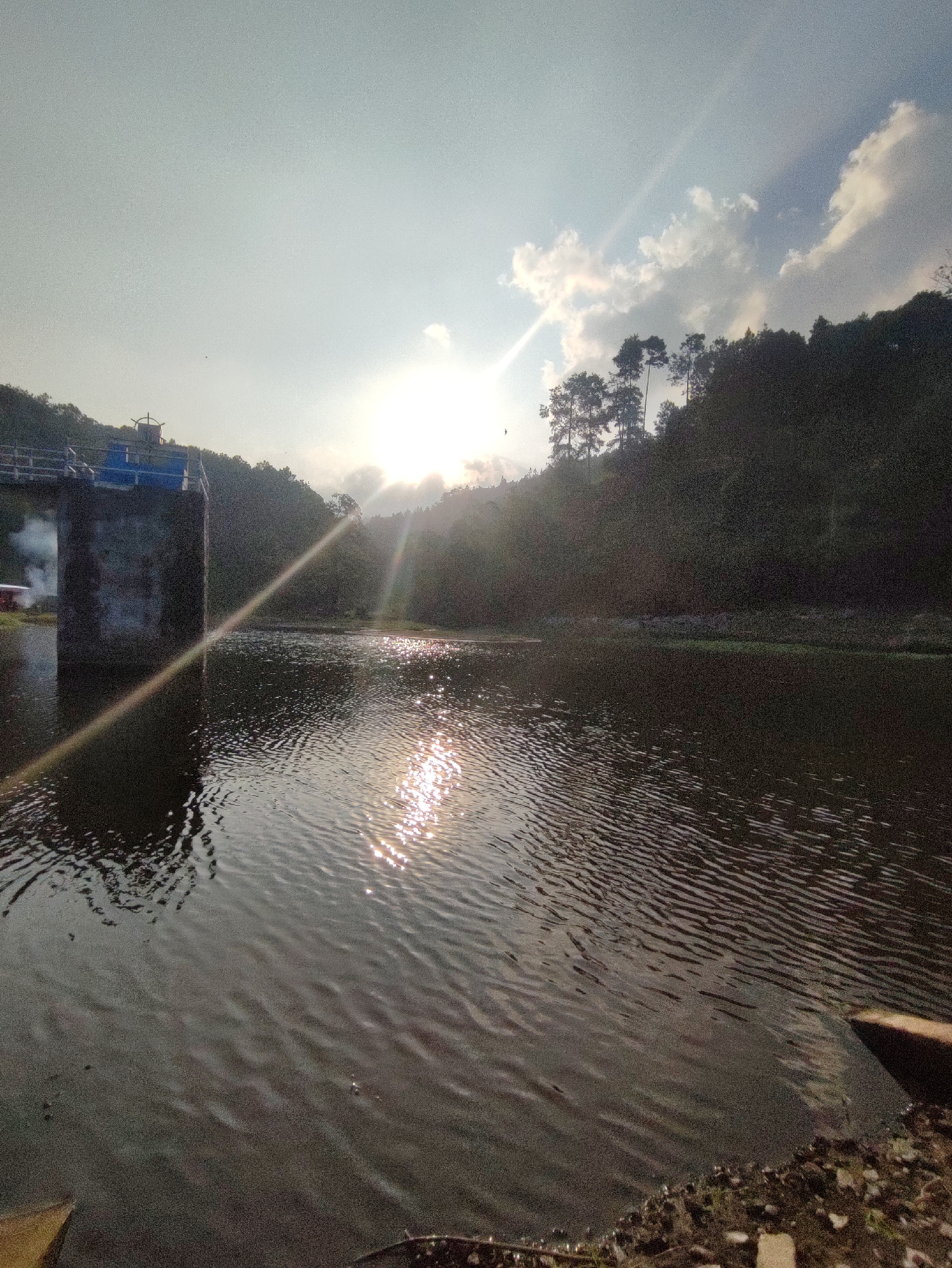 The image shows a body of water in front of a concrete structure that appears to be part of a dam or water control system. The sun is setting or rising, casting a bright glare on the water and creating light rays that extend across the scene. The background consists of a lush, tree-covered hillside. The overall impression is a serene natural landscape with man-made elements. The location is identified as Joho, Indonesia, a city known for its natural beauty and rural charm.