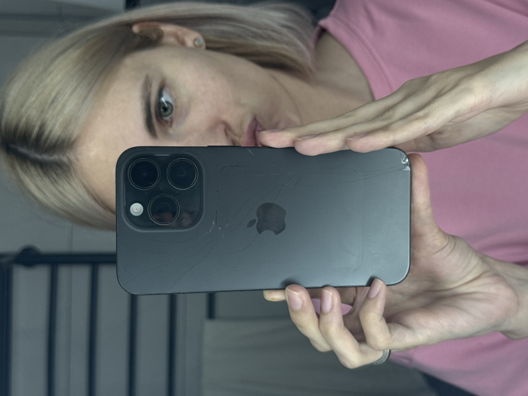 A woman is holding a cracked iPhone to take a selfie in a mirror. The phone is dark gray and has three camera lenses. Cracks are visible on the back of the phone around the Apple logo. The woman has fair skin, blonde hair, and is wearing a pink shirt. The background is blurry, but it appears to be an interior space with vertical lines. The location in the city of Saratov, Russia cannot be verified.