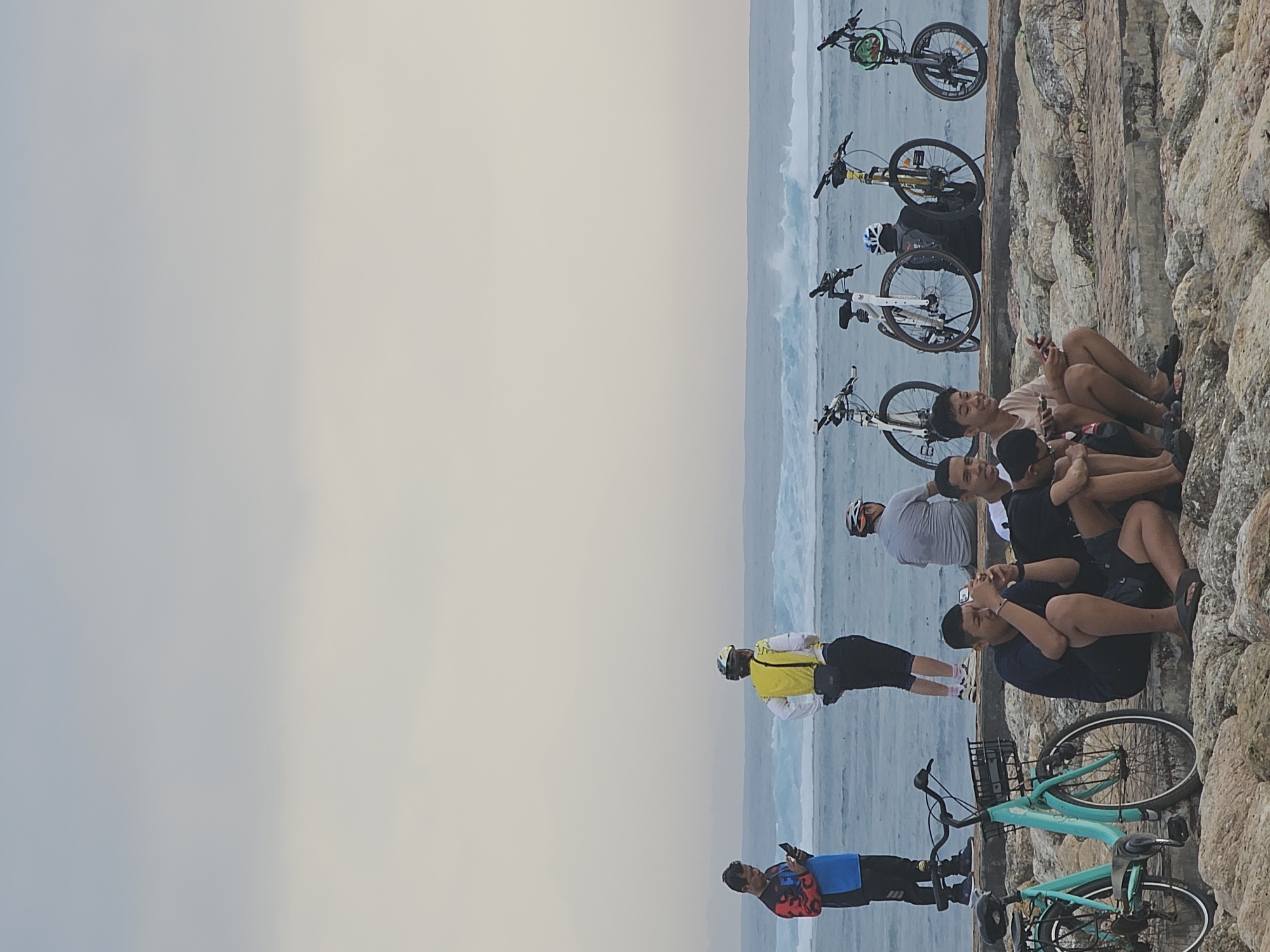 The image depicts a group of people and several bicycles on a rocky embankment with the ocean and waves in the background. The sky is overcast, suggesting it might be dawn or dusk.
In the foreground, several individuals are seated on the rocks. One young man in a dark blue t-shirt and shorts is looking at his phone. Next to him, another young man is looking at his phone, and to his right, a third young man is also looking at his phone. Further to the right, another person is seated, partially obscured.
Further back, standing on the embankment, are two individuals. One person on the left is wearing a blue and red long-sleeved shirt and black pants, holding a phone. To their right, another person is wearing a yellow vest over a white shirt and black pants, facing away from the camera. Behind them, a person in a grey shirt is seated, and a bicycle is parked nearby.
Several bicycles are positioned on the embankment. Two bicycles are visible on the left, one teal and one white. Further to the right, there are three more bicycles, two white and one yellow, parked in a row. The bicycles appear to be for rent, with some having baskets.
The environment includes a rocky surface in the foreground, transitioning to a paved embankment, and then the ocean with waves. The location is likely a coastal area in Denpasar, Indonesia, possibly a beach or promenade known for cycling and ocean views.
