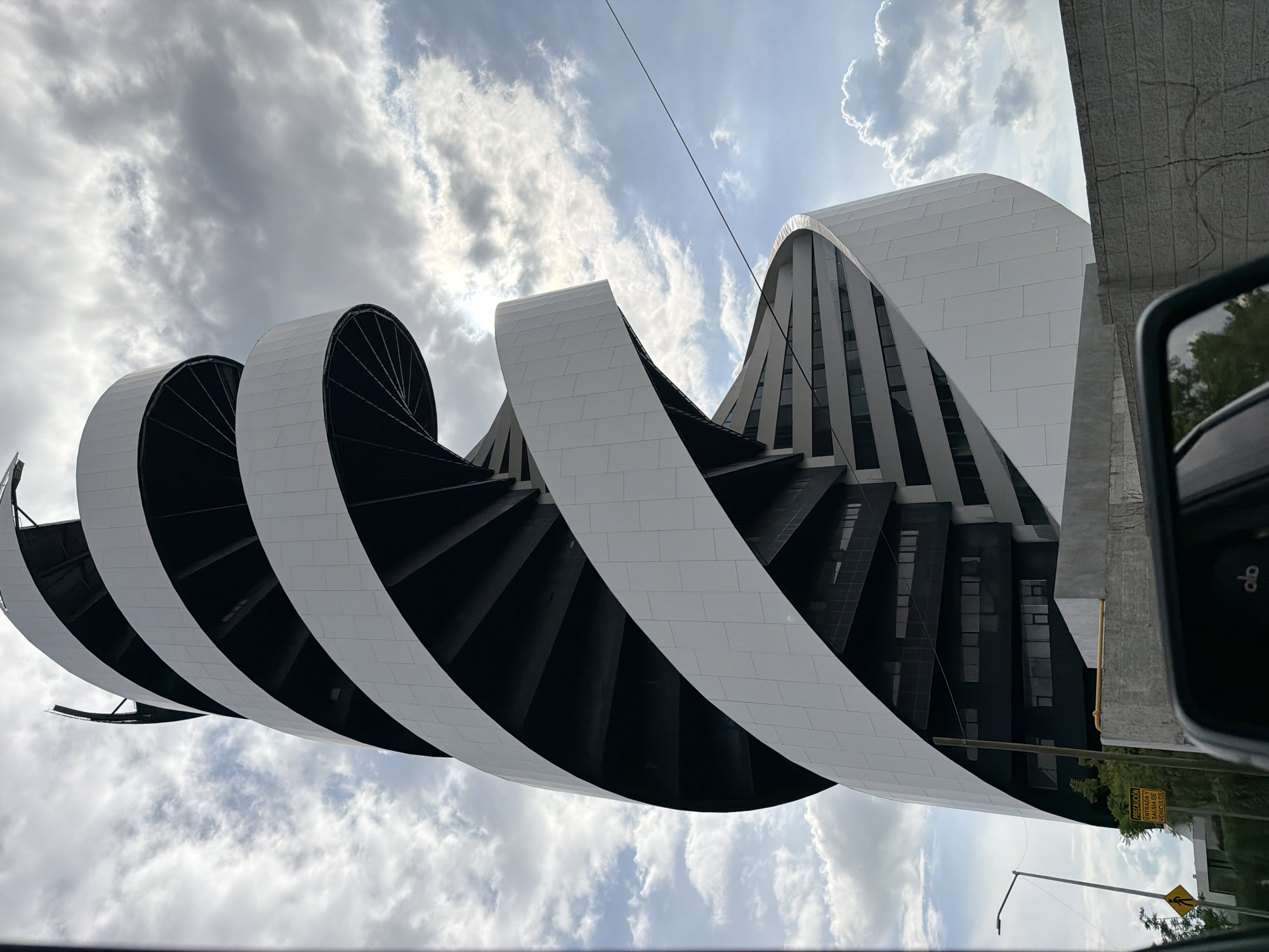 The image captures a striking, modern building with a unique spiral design, located in San Bernardino Tlaxcalancingo, Mexico. The building's exterior is predominantly white, with black accents visible between the spiraling levels, suggesting floors or open spaces. The structure tapers as it ascends, creating a dynamic, twisting silhouette against a cloudy sky. In the foreground, a section of a car's interior, including a side-view mirror, is visible, indicating the photo was taken from a vehicle. Beyond the building, a street lamp and a yellow warning sign are present. The overall scene is urban, with the architectural marvel serving as the central focus.