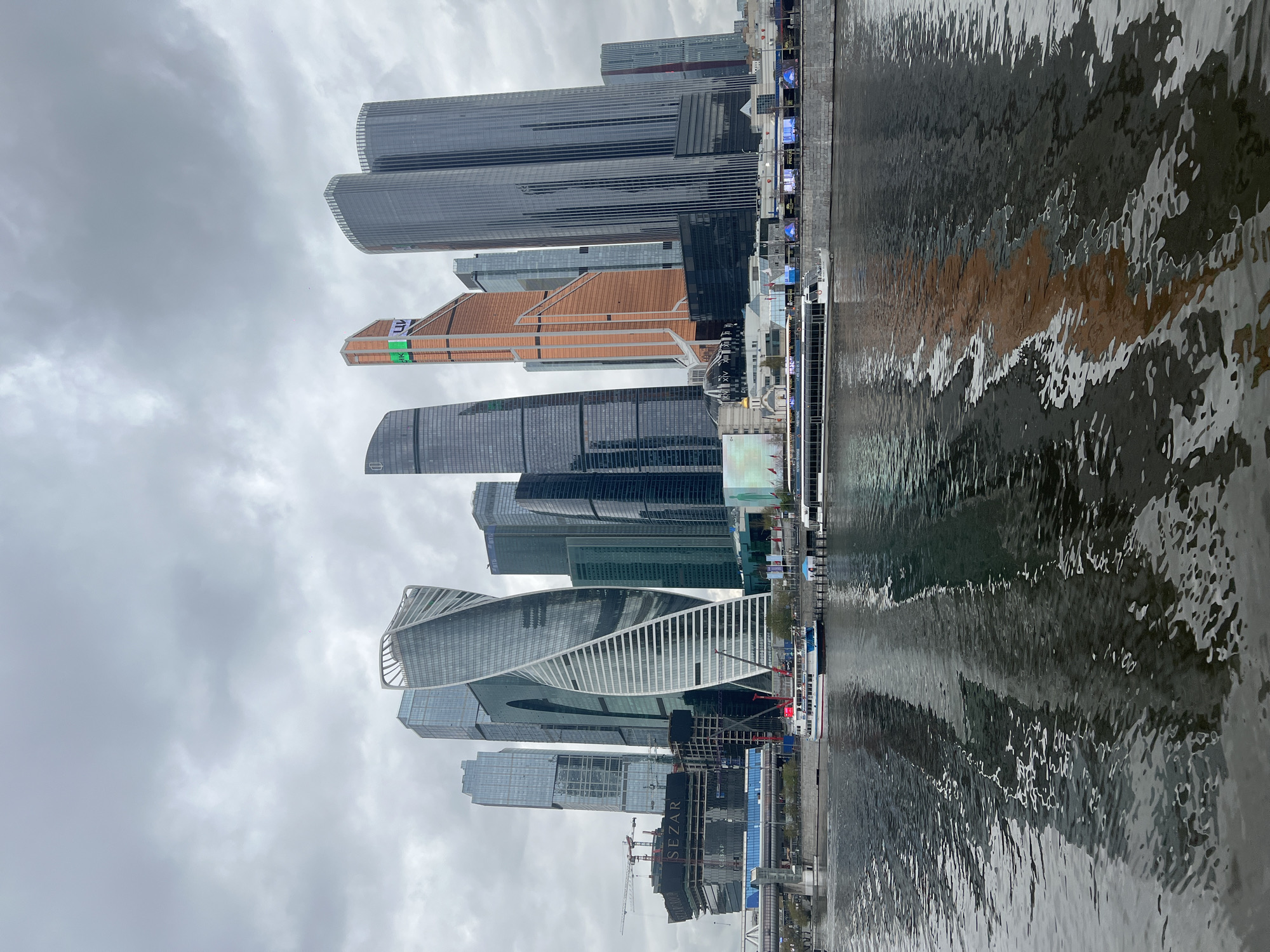 The image shows the Moscow International Business Center (Moscow City) in Khimki, Russia, reflected in the water. Several skyscrapers are visible, including the Evolution Tower with its distinctive twisted shape. Other buildings of varying heights and architectural styles stand alongside. The sky is overcast with gray clouds. Water fills the lower portion of the image, creating a distorted reflection of the city skyline. Construction cranes and other construction materials are visible near the base of the buildings along the water's edge. A passenger boat is docked along the river's edge.