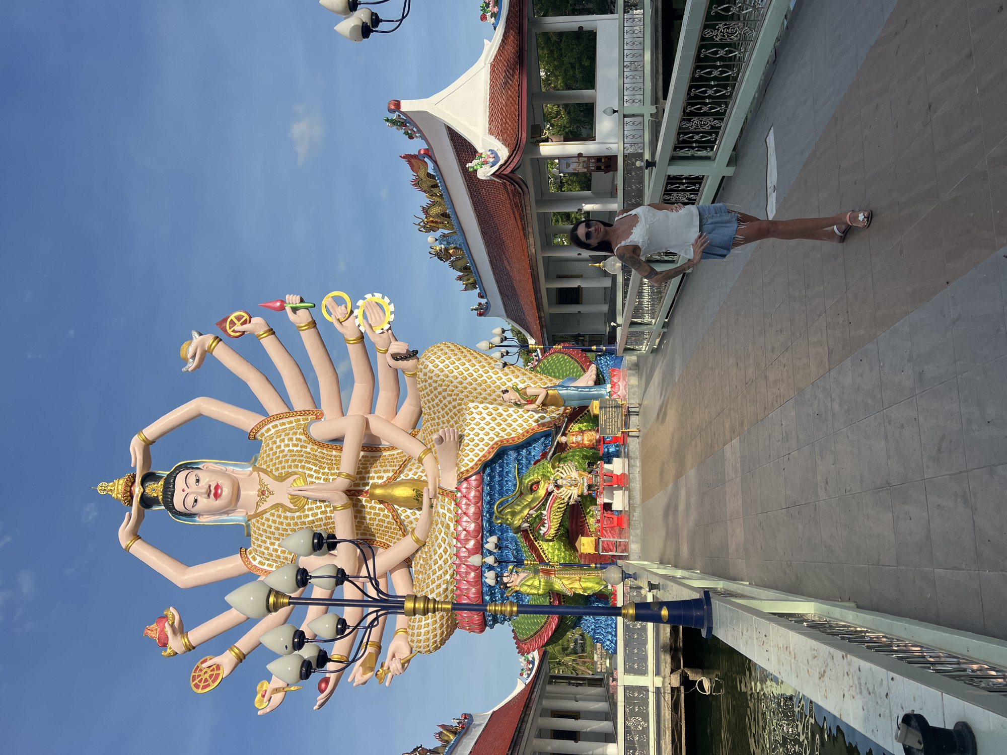 A woman stands on a bridge in Baan Plai Laem, Thailand, next to a large, ornate statue with multiple arms. The statue is adorned with golden embellishments and rests upon a colorful base featuring a green dragon. The bridge has a patterned railing and tiled surface. A traditional Thai-style building with a red and white roof is visible behind the woman. The sky is clear and blue.