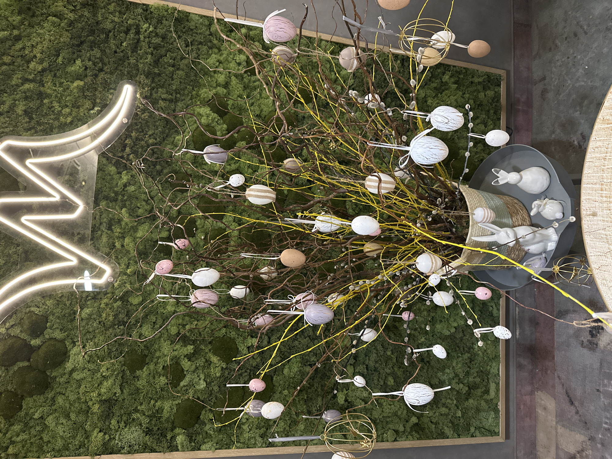 A high-angle, indoor shot depicts an Easter-themed display, possibly in Wrocław, Poland. At the center is a dense bouquet of brown and yellow branches adorned with pastel-colored Easter eggs, placed in a basket resting inside a gray circular container that holds ceramic rabbit figurines. The background features a wall covered in green moss with a neon lightning bolt sign attached to it. The floor on the right side is gray and has some paint on it.