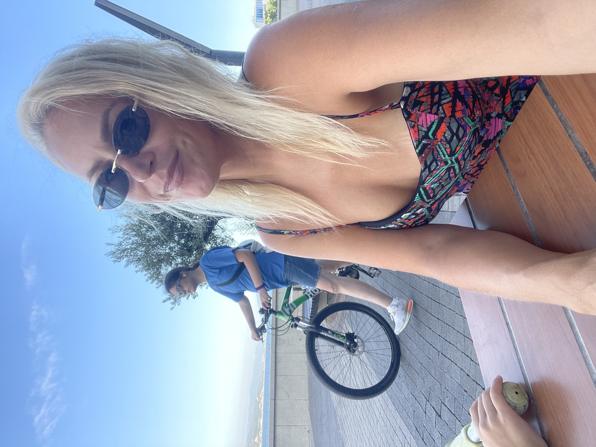 The image shows a woman with blonde hair and sunglasses in the foreground, smiling at the camera. She is wearing a patterned dress and sitting at a wooden table. In the background, another person is riding a bicycle along a paved path next to a body of water. The sky is blue with a few clouds. The location is the city of Ivanovskaya, Russia.