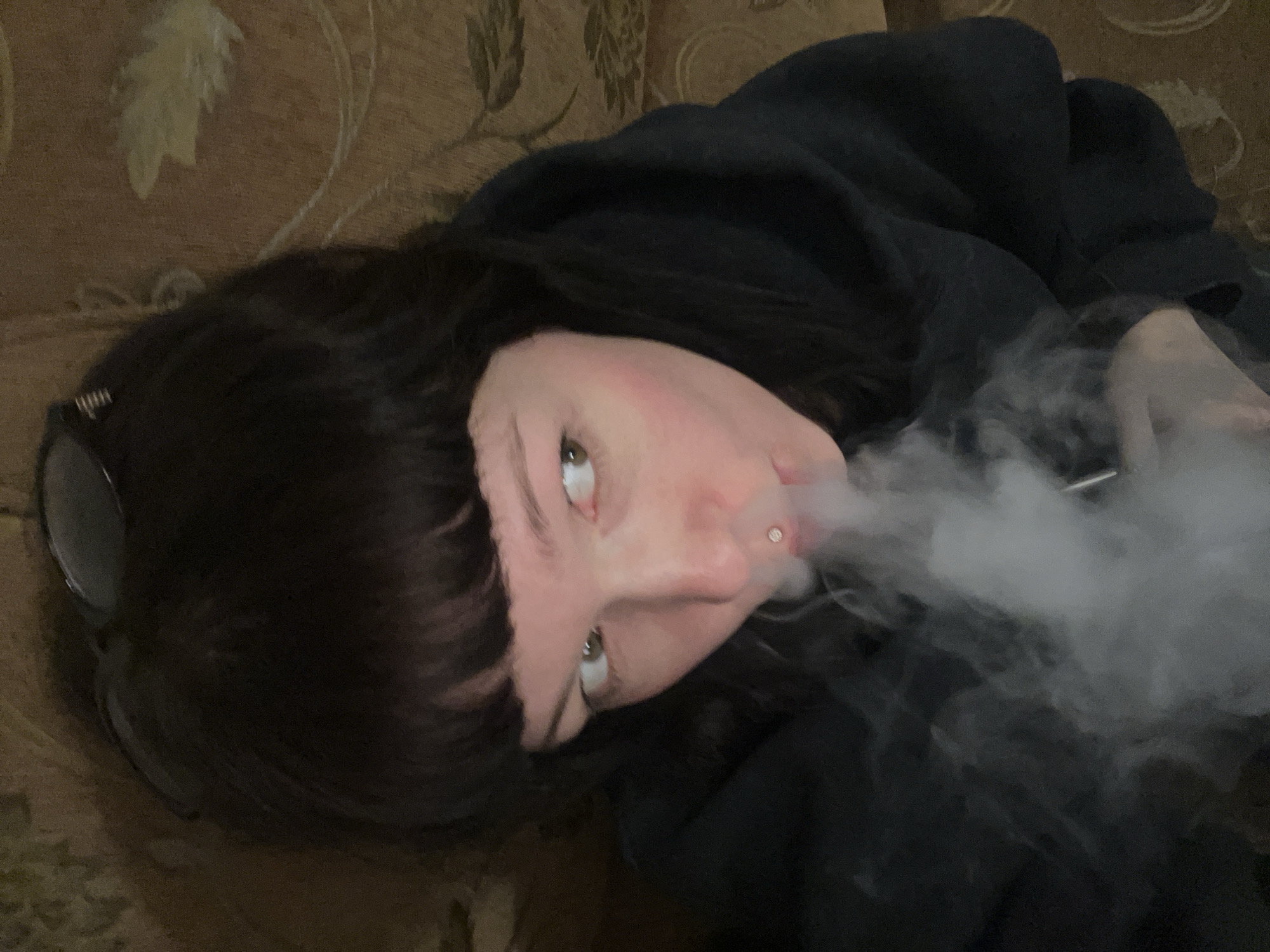 A person with dark hair and bangs is lying on a patterned couch, exhaling a large cloud of vapor. Glasses are resting on their head. A piercing is visible on their lip. The person is wearing a dark hooded sweatshirt. The location is unknown.