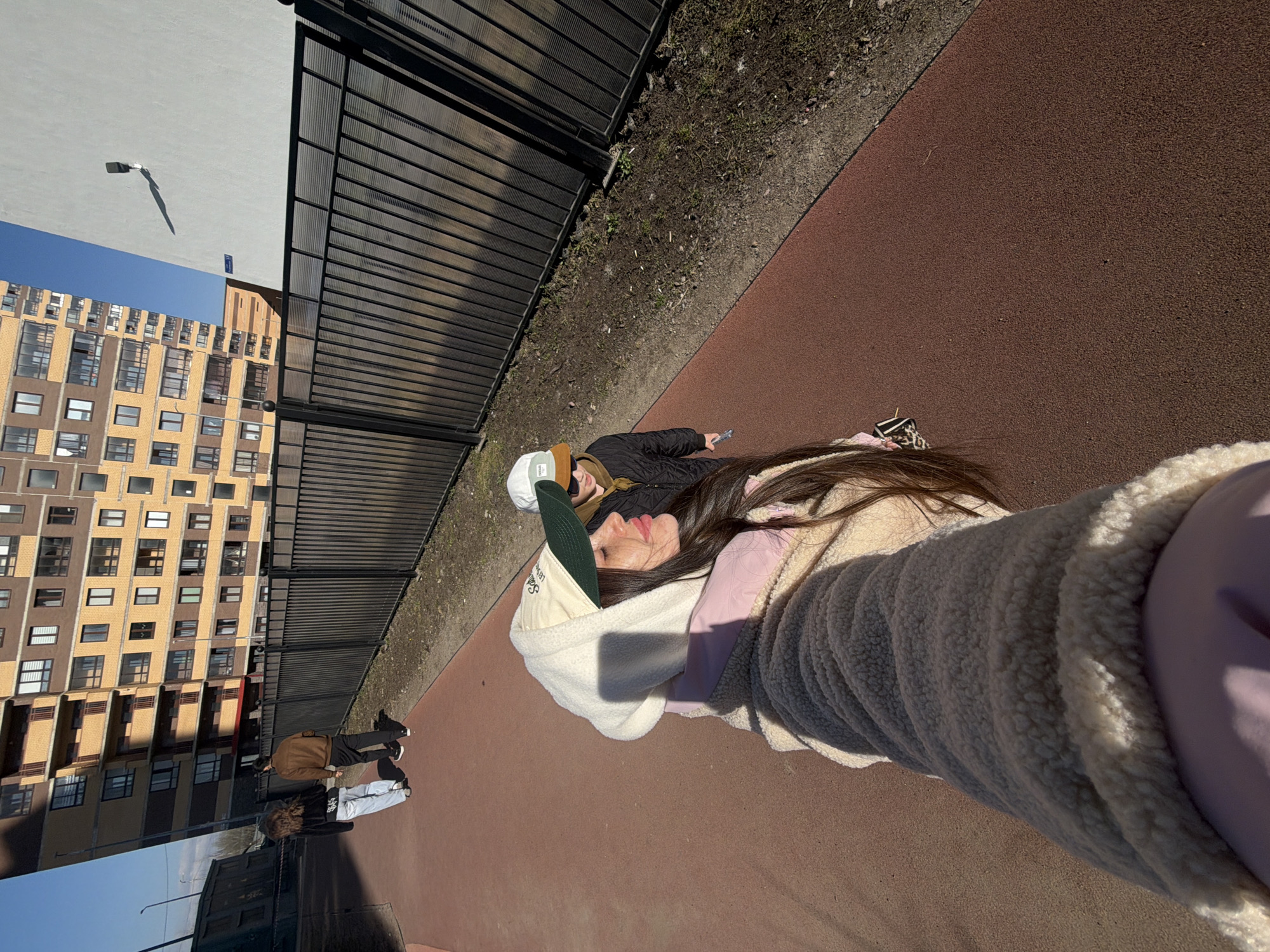 The image shows a woman taking a selfie outdoors. The woman is wearing a beige sherpa-lined hoodie over a lavender shirt and a green and white baseball cap. A person is lying next to her with a jacket on. A black fence separates them from a narrow strip of soil, and then a red walking surface. In the background, there's a multi-story apartment building with a beige facade and numerous windows. The weather appears sunny. Based on visual evidence, the scene could be located in Saint Petersburg, Russia.