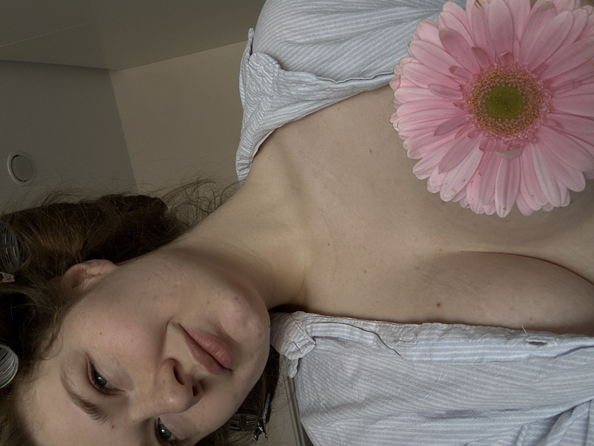 The image depicts a person lying down, possibly indoors, with their head tilted towards the lower left corner of the frame. They have fair skin, light-colored eyes, and brown hair with curlers visible near the top of the head. The person is wearing a light blue and white striped shirt. A large pink gerbera daisy is positioned on their chest, obscuring part of the shirt. The background is out of focus, showing a light-colored wall and ceiling. The setting appears to be indoors, potentially a bedroom or bathroom. The image does not provide enough information to determine the location as Kazan, Russia.