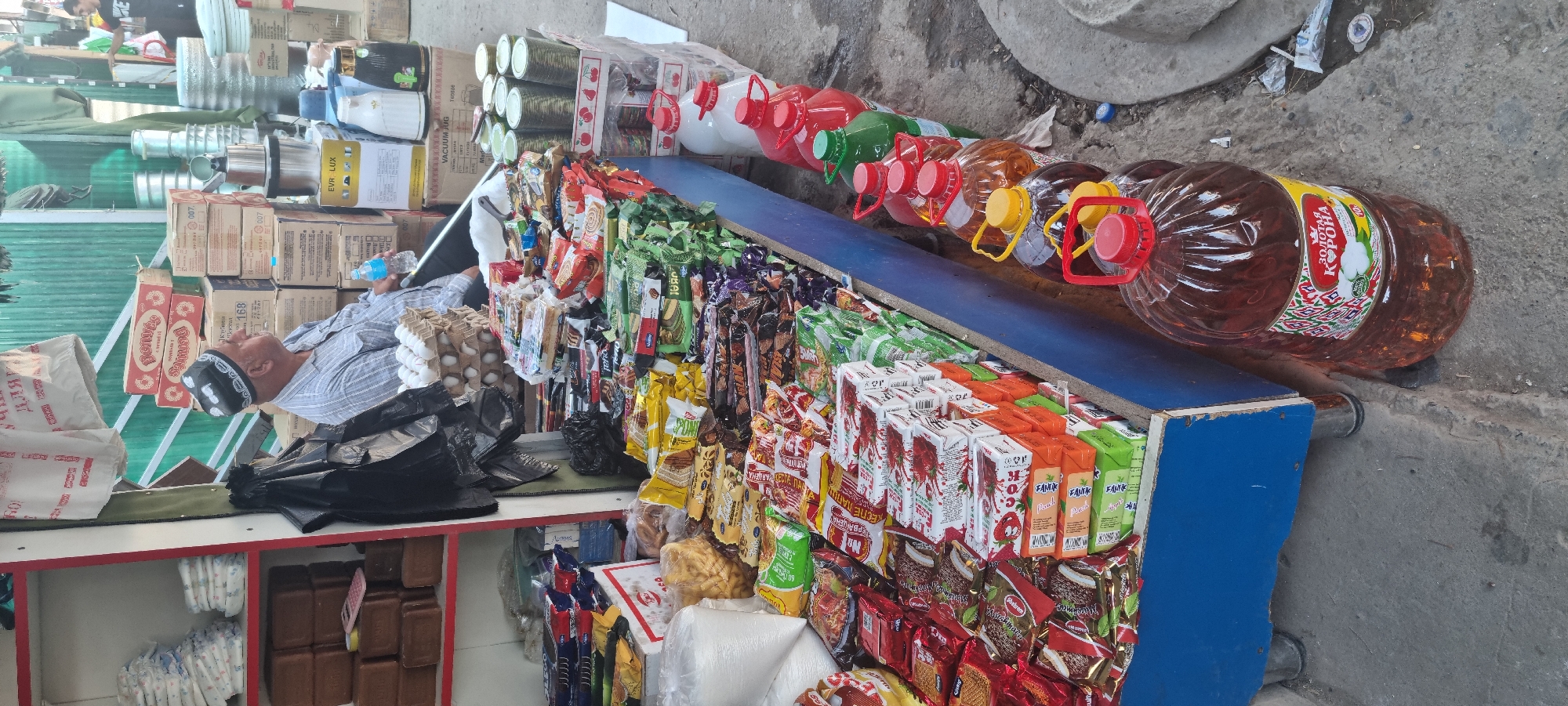 The image depicts an outdoor market stall in Bokhtar, Tajikistan. The stall is laden with various goods, primarily packaged food items like crackers, cookies, and instant noodles, displayed in colorful wrappers and boxes. A man wearing a traditional skullcap and a striped shirt sits behind the stall, holding a bottle of water. In front of the stall, there are several large plastic bottles filled with a dark amber liquid, likely cooking oil, arranged in a line. Behind the man, stacked boxes and metal containers are visible. The ground is paved, and the stall appears to be a permanent structure with a blue-painted wooden counter and metal legs. The overall impression is of a bustling local marketplace.