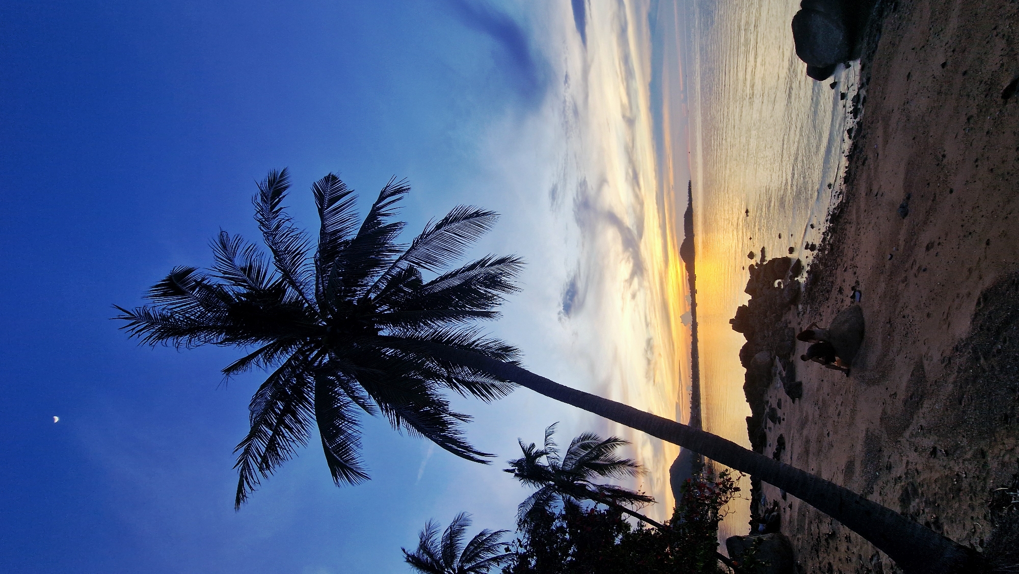 A palm tree is silhouetted against a vibrant twilight sky over the ocean in Baan Bang Rak, Thailand. The sky transitions from deep blue at the top to lighter hues of orange and yellow near the horizon where the sun has set. A crescent moon is visible in the upper left quadrant of the sky. The foreground consists of a sandy beach with scattered rocks and a few dark boulders. In the middle ground, a couple is seated on a large rock on the beach. The ocean is calm, reflecting the colors of the sky. In the distance, islands and a coastline are faintly visible. Other palm trees and foliage are silhouetted on the left side of the image.