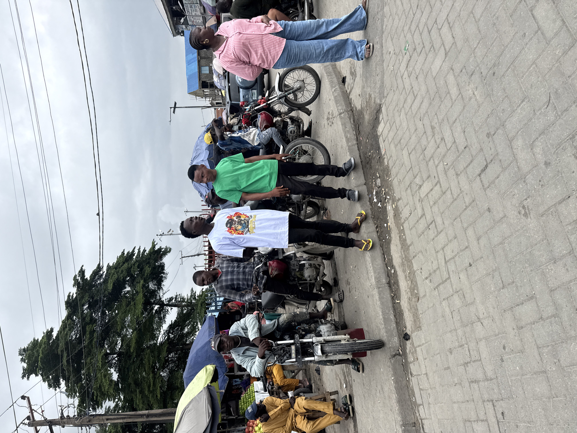 The image depicts a street scene in Ado, Nigeria, likely a market area, characterized by a gray, overcast sky and a paved walkway. In the foreground, four young men stand near several motorcycles. The man on the far left wears a light blue jacket and sits on a motorcycle, looking forward. To his right, another man in a plaid shirt stands beside a motorcycle. A third man, in a white t-shirt with a graphic print, stands next to him, also looking forward. The fourth man, wearing a bright green t-shirt, stands to the left of the man in the white t-shirt. Further back, other individuals and vehicles are visible. A woman in a pink and white striped shirt and blue jeans stands on the right side of the image, facing away from the camera. Market stalls with umbrellas are set up along the side of the street, with produce visible in one stall. The environment suggests an active commercial zone with transportation and pedestrian activity.