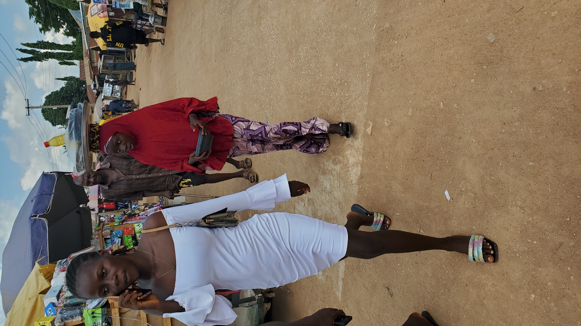 A medium shot captures a bustling street scene in Jalingo, Nigeria. In the foreground, a young woman, dressed in a white off-the-shoulder dress with bell sleeves, walks while talking on her phone. Her right hand is raised to her ear, and a patterned handbag hangs from her left shoulder. She wears colorful, jeweled slide sandals. To her right and slightly behind her, a young girl in a red hijab and patterned trousers walks, carrying a dark rectangular object in her hands. She has items balanced on her head, including a yellow bottle and two round, flat objects on a metal tray. Between them, an older man wearing a brown checkered shirt and dark pants walks with a cane, looking towards the right. The background reveals a market setting with stalls and vendors, obscured by an umbrella. A power pole and trees are visible further in the distance. The ground is dusty and uneven.