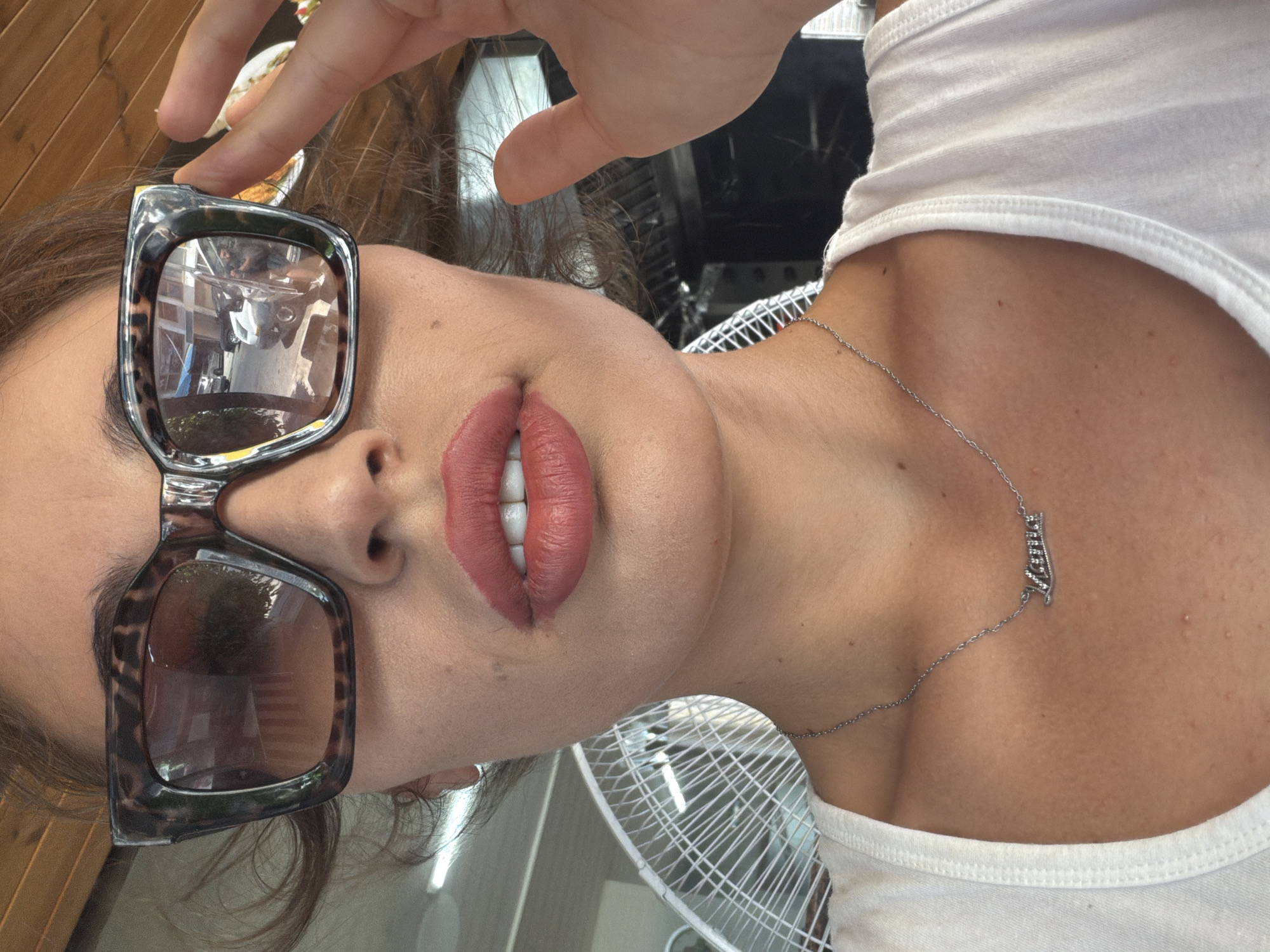 A close-up selfie shows a woman's face from the nose down, as she adjusts her large, leopard-print sunglasses. Her lips are painted a matte coral-red, and she wears a delicate silver necklace with the name "Larna" in cursive. Behind her, a white oscillating fan is visible, along with the blurred background of what appears to be a restaurant or cafe. The reflection in her sunglasses shows a street scene with cars and buildings, indicating she is likely in a city setting, possibly Larnaca, Cyprus, given the name on her necklace. The woman is wearing a white tank top.