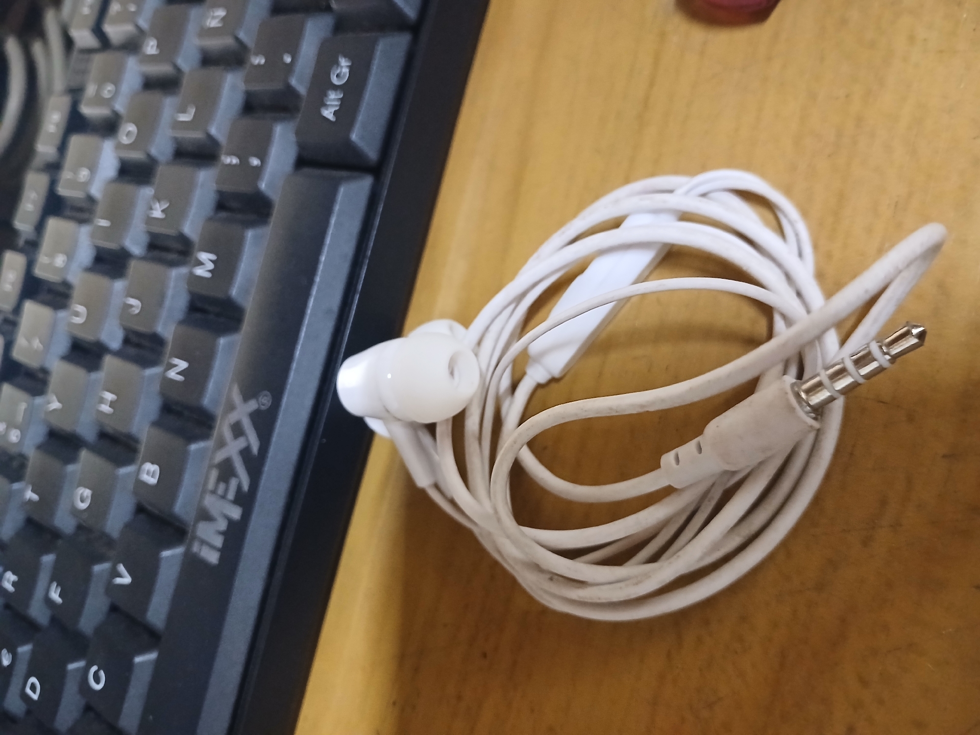 A close-up, slightly angled shot shows a white wired headset tangled on a wooden surface, next to the edge of a black keyboard. The headset has two white earbuds and a white inline microphone with a single button. The cable connects to a silver 3.5mm audio jack with three bands. The keyboard is partially visible on the left side of the frame, with keys showing letters such as "Alt Gr", "M", "H", "G", "B", "F", "R", "C", and "O". The text "iMEXX" is visible on the keyboard. The lighting suggests an indoor setting, likely during the day. The overall scene is that of a personal workspace, with everyday technology items arranged on a desk.
