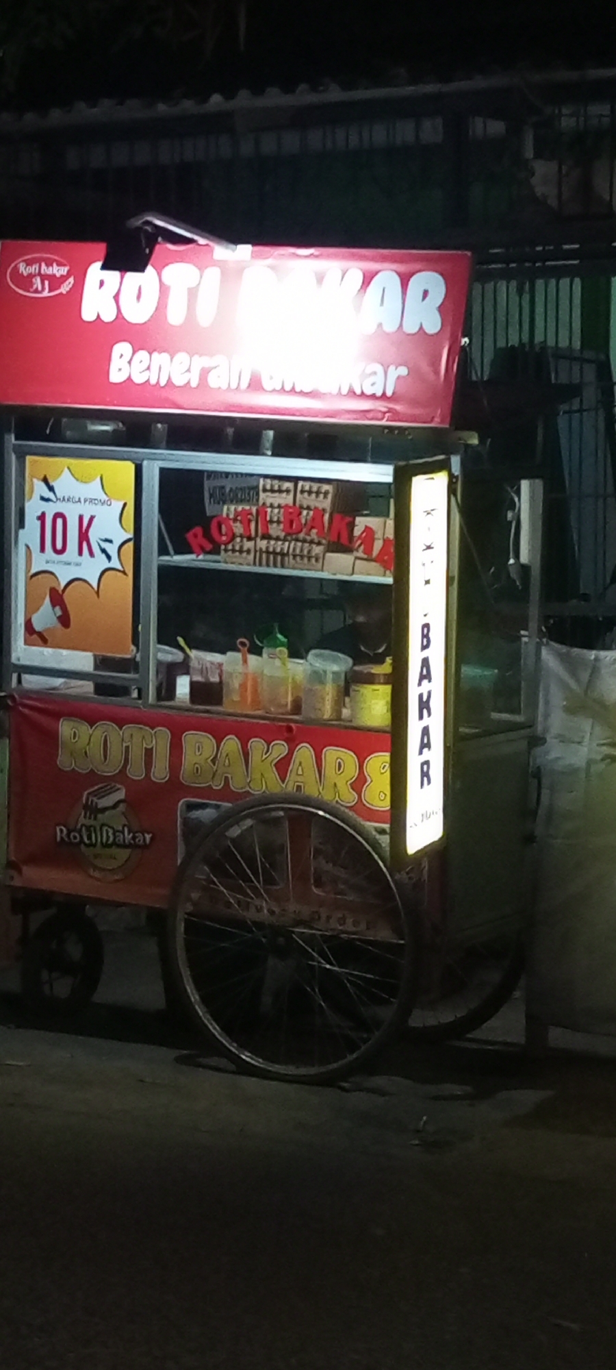 An outdoor night shot captures a "Roti Bakar" (toasted bread) food cart. The cart is red with white lettering. The sign at the top reads "ROTI BAKAR" in large white letters, with a smaller line below that says "Beneran Bakar". To the left of the main sign, there is a smaller logo with text that says "Roti Bakar". On the left side of the cart, a yellow sign with a red megaphone and the text "HARGA PROMO 10 K" is displayed. Inside the cart, shelves display bread and various condiments in jars and bottles. A vertical illuminated sign on the right side of the cart displays the text "BAKAR" vertically. Two large spoked wheels are visible at the bottom of the cart. The cart appears to be parked on a street in Setu, Indonesia.