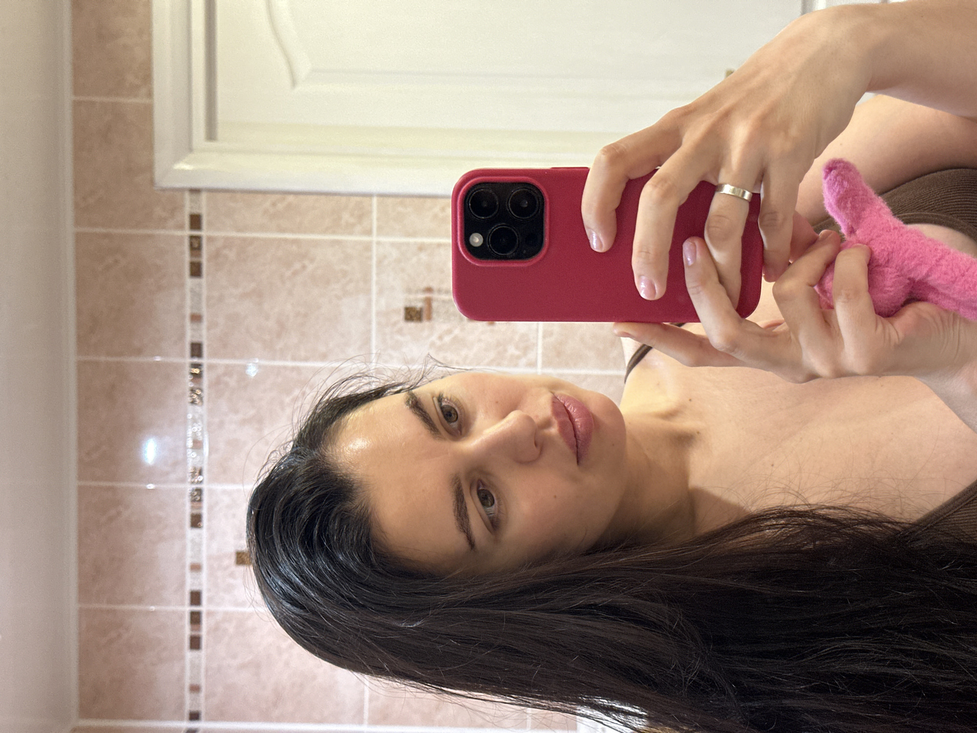 A mirror selfie features a woman with long, dark hair and a neutral expression, looking directly into the camera. She holds a red-cased smartphone, likely an iPhone due to its visible triple camera system, in her right hand. A silver ring is worn on her right ring finger, and her fingernails have a light polish. Her left hand is partially visible, holding a pink, fluffy object, which appears to be a small towel or cloth. She is wearing a dark-colored top or bra.
The background, reflected in the mirror, reveals an indoor setting, likely a bathroom. The wall is covered with light brown or beige square tiles, interspersed with a vertical decorative strip of smaller, darker, and slightly reflective tiles. Above the tiled section, a white door or cabinet with panel detailing is visible. The overall lighting is soft and even. No text is visible in the image. The location context for this image is Chaikovsky, Russia.