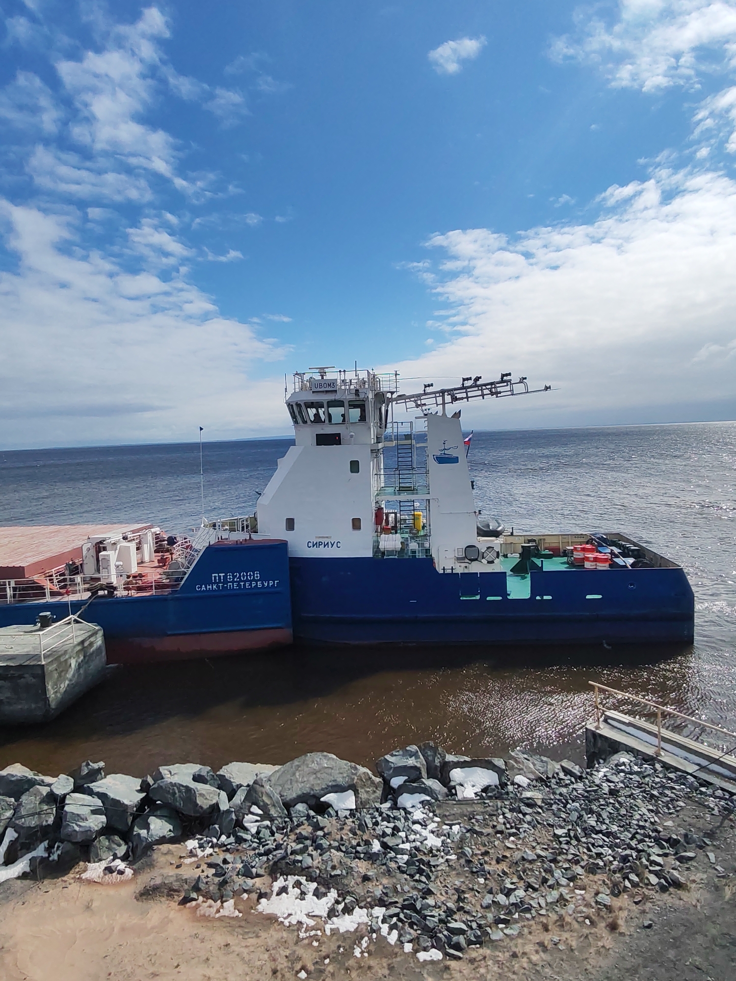 A large blue and white workboat, possibly a tugboat or icebreaker, is docked alongside a rocky and gravelly shore under a bright blue sky with scattered white clouds. The vessel, named "СИРИУС" (Sirius) on its superstructure, features a prominent bridge with various antennae and equipment. Below the name, the hull displays "ПТ82008" and "САНКТ-ПЕТЕРБУРГ" (Saint Petersburg), indicating its registration and home port. A Russian flag is visible on the stern.
The vessel is connected to a smaller, reddish-brown barge or another section of a vessel on its port side, which is partially obscured. On the shore, there are large grey rocks, smaller gravel, and patches of melting snow or ice, suggesting a cold climate or season. The water near the shore is a murky brown, transitioning to a dark blue further out towards the horizon. On the right side of the frame, a small ramp or jetty with a railing extends into the water from the shore. No people are visible. The scene depicts a functional maritime environment, likely in Shcheleiki, Russia, during the daytime with clear weather.
