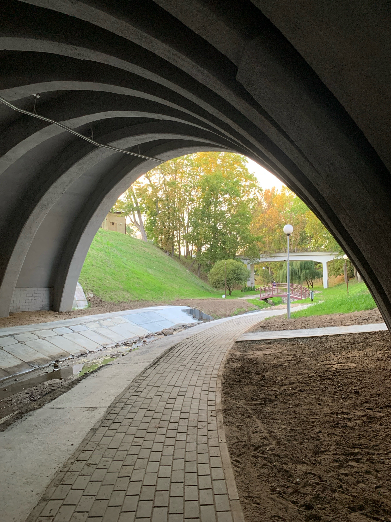 The image shows a pathway under an arched concrete structure. The pathway is paved with light-colored rectangular bricks and curves to the right. To the left of the pathway, there is a concrete drainage channel with a small amount of water and debris. Beyond the drainage channel, there is a grassy slope leading up to trees. In the distance, a small pink bridge crosses a body of water, and further beyond that, a white pedestrian overpass spans across the scene. Two lampposts are visible, one on the right side of the pathway and another in the middle ground near the pink bridge. The ground to the right of the pathway is dark soil with visible tire tracks. The overall environment appears to be a park or recreational area in the city of Hrodna, Belarus.