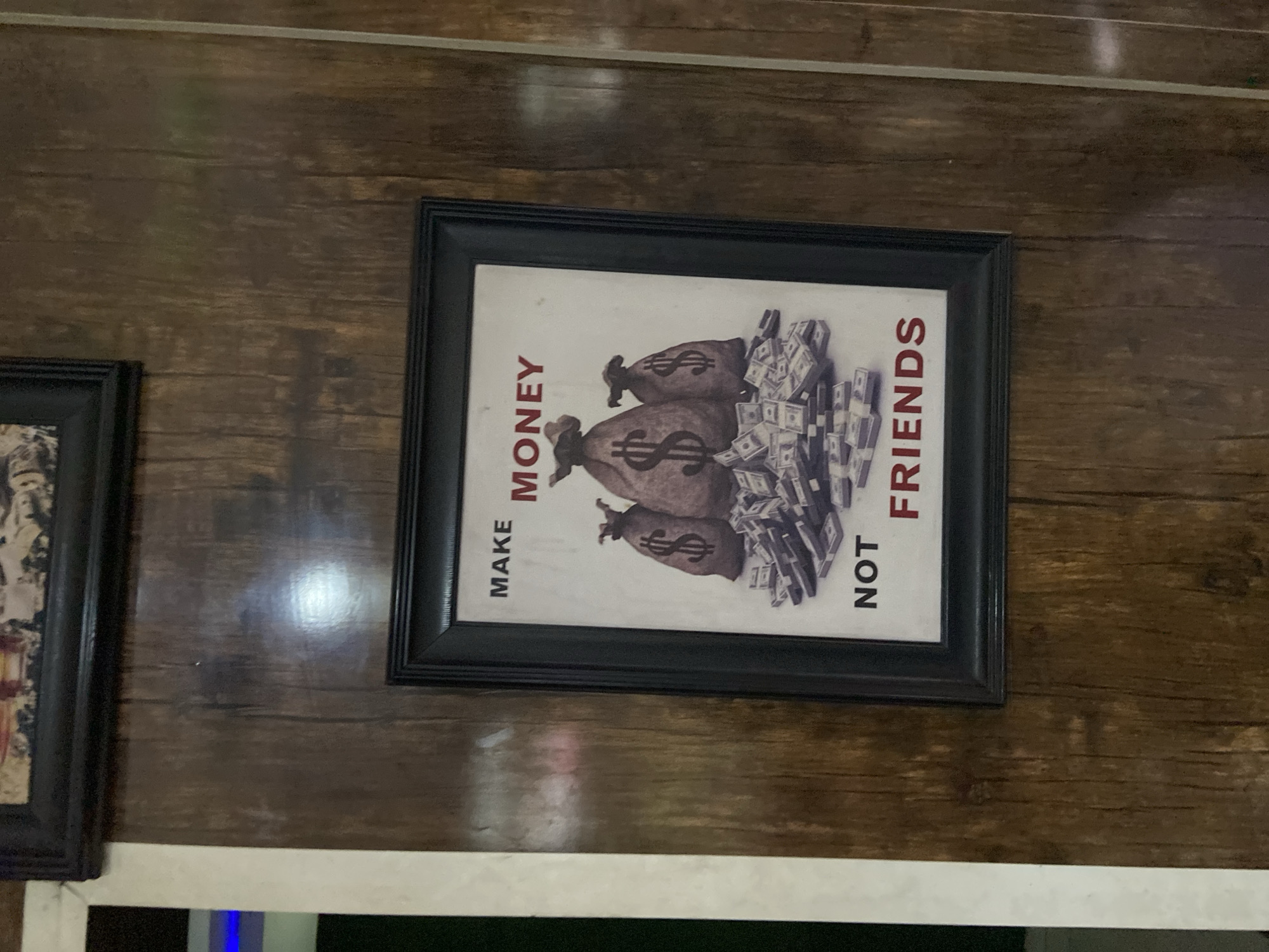 A framed picture hangs on a brown wooden wall. The picture contains an image of money bags with dollar signs on them and stacks of money. Written vertically on the left of the picture is the phrase "MAKE MONEY" and on the right is the phrase "NOT FRIENDS". To the left of the framed picture is another picture frame. The environment is indoors. The city and country are not ascertainable from the image.