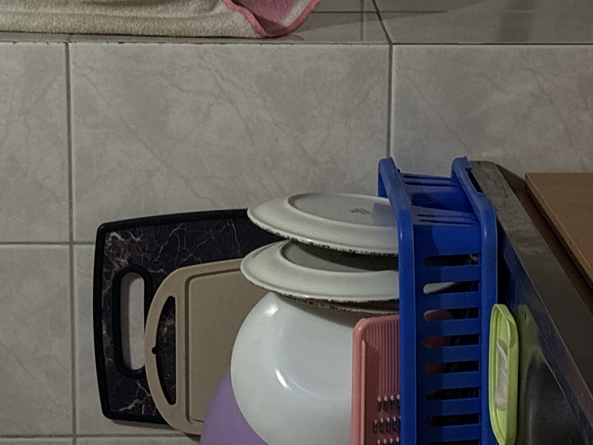 A close-up shot depicts a kitchen setting in Murino, Russia. The tiled wall in the background features light grey tiles. A kitchen dish rack in blue is positioned on a dark surface. Inside the rack, there are two white plates stacked on top of each other. To the left of the plates are two cutting boards: one black with a marbled pattern and one beige with a handle. There is a white bowl visible below the plates, and a pink grater is partially visible. Additionally, a green scrubber is attached to the side of the blue dish rack. A beige towel with a pink edge is seen at the top of the frame.