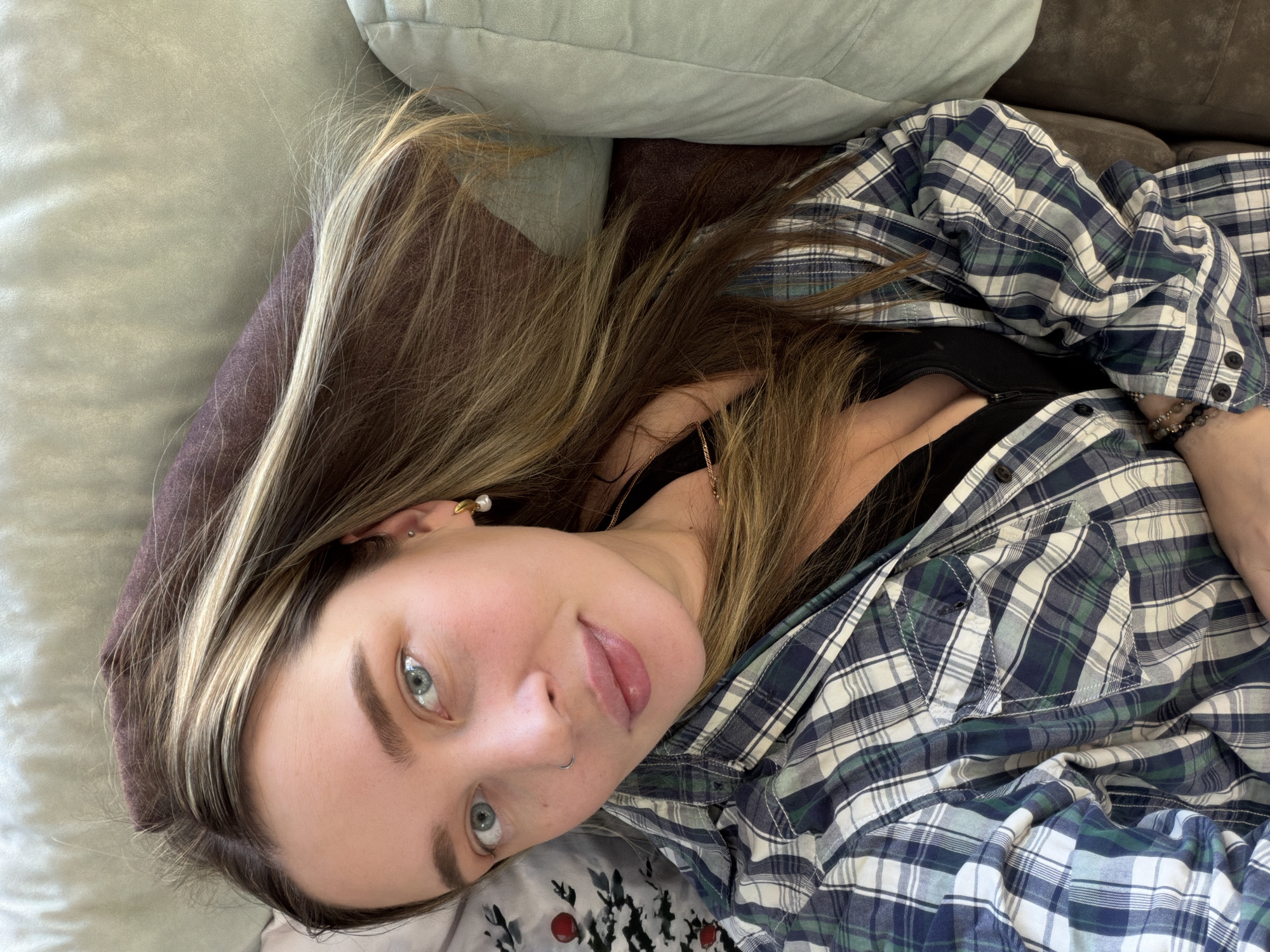A young woman with long, light brown hair and blonde highlights lies down, gazing directly at the camera. Her head is tilted, presenting a diagonal composition. She has striking light blue or grey eyes, neatly groomed eyebrows, and fair skin. A small septum piercing adorns her nose, and a gold-colored earring with a pearl drop is visible in her left ear. Her lips are slightly parted, conveying a relaxed expression.
She is wearing a blue, green, and white plaid button-up shirt, unbuttoned to reveal a black top or undergarment beneath. A delicate gold-toned necklace is visible around her neck, and a beaded bracelet is on her left wrist. She is positioned on what appears to be a couch or bed, with a light grey-green pillow or cushion behind her and a dark brown surface. Part of a patterned fabric, possibly a pillowcase or blanket featuring red berry-like designs and dark leaves, is visible below her head.
The setting is an indoor, casual environment, likely a home in Chaikovsky, Russia. The lighting is soft and ambient, suggesting a relaxed moment without specific time-of-day indicators. No text is visible in the image.
