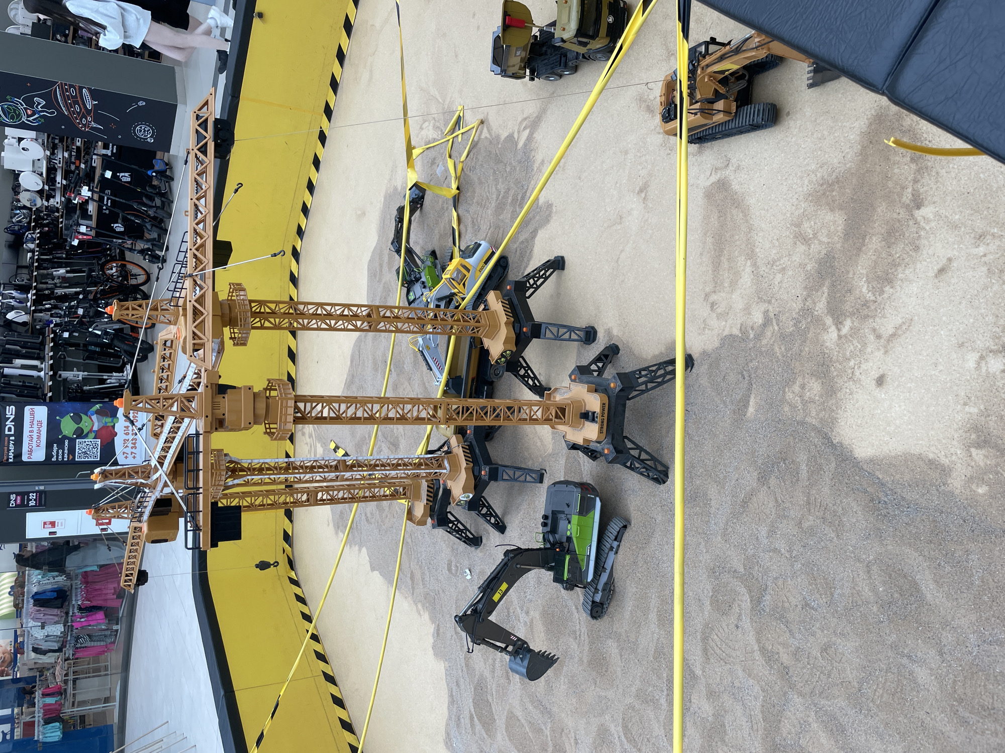 The image depicts an indoor sandbox play area, possibly at a children's entertainment center or a store. Several large, toy construction vehicles, including excavators and dump trucks, are arranged on the sand. Towering over the vehicles are several large, toy construction cranes, their yellow booms extended over the sandpit. Yellow caution tape demarcates the perimeter of the sandbox, with black and yellow hazard stripes on the surrounding wall. In the background, a retail environment is visible, with racks of clothing and displays of electronic goods, suggesting it is located within a shopping mall or department store. Two young women are walking in the background, one in a light-colored sweatshirt and skirt, the other in a dark outfit. The scene is well-lit, and the sand appears to be clean. There are no obvious signs of damage or distress. The location is identified as a shopping mall in Perm, Russia.