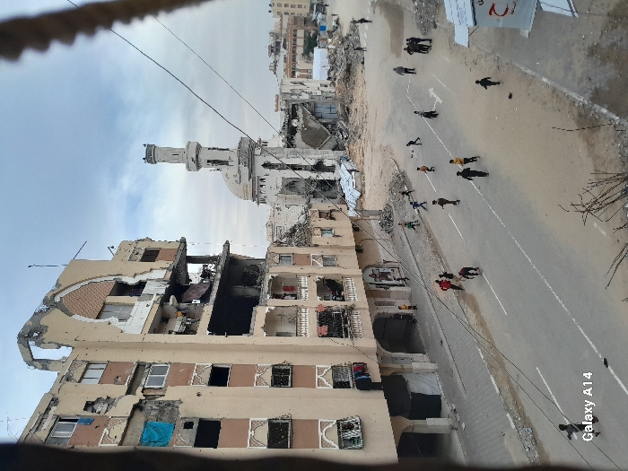The image depicts an urban street in Khan Yunis, Palestinian Territory, marked by extensive structural damage under an overcast sky. To the left, a multi-story, light-colored building exhibits severe damage, with large sections of its facade missing, exposing interiors. Bullet or shrapnel impacts are visible across its surface, and some windows are covered with blue tarpaulins. Laundry hangs from balconies on lower floors. In the background, to the right, the prominent minaret and dome of a mosque are heavily damaged, surrounded by debris, indicating significant structural collapse.
A wide, paved road with faded white lane markings (a double solid line and a single solid line, along with a partial arrow) spans the mid-ground. Approximately 15 to 20 children, casually dressed, are scattered across the street, appearing to play or run. On the far right side of the street, a small group of 3-4 adults stands near a debris-strewn area. Piles of rubble and demolished structures are visible both adjacent to the street and in the background, especially surrounding the mosque. Utility wires are strung horizontally across the mid-air. A small kiosk-like structure with partial Arabic text is present on the right edge of the foreground. The scene conveys widespread urban destruction.