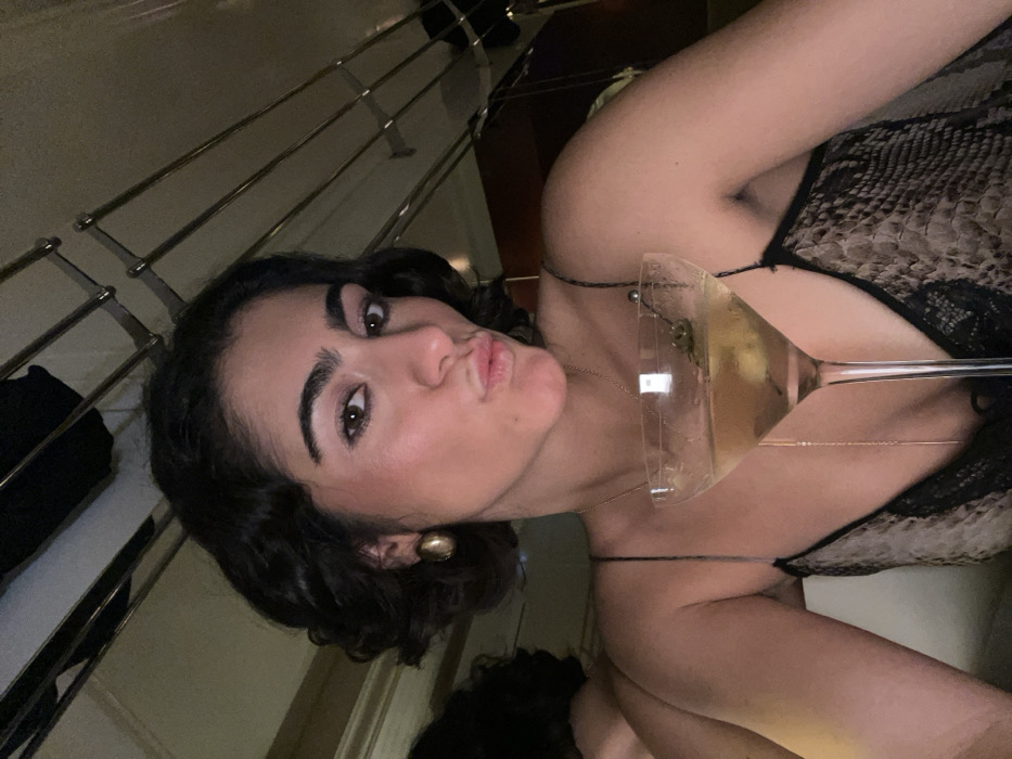 A woman with dark curly hair poses close to the camera with a pout, holding a martini glass with a golden liquid inside. She wears a snakeskin print top with black lace details and thin spaghetti straps. A gold necklace is visible. The background features a metal railing and dark furniture, suggesting an indoor setting.