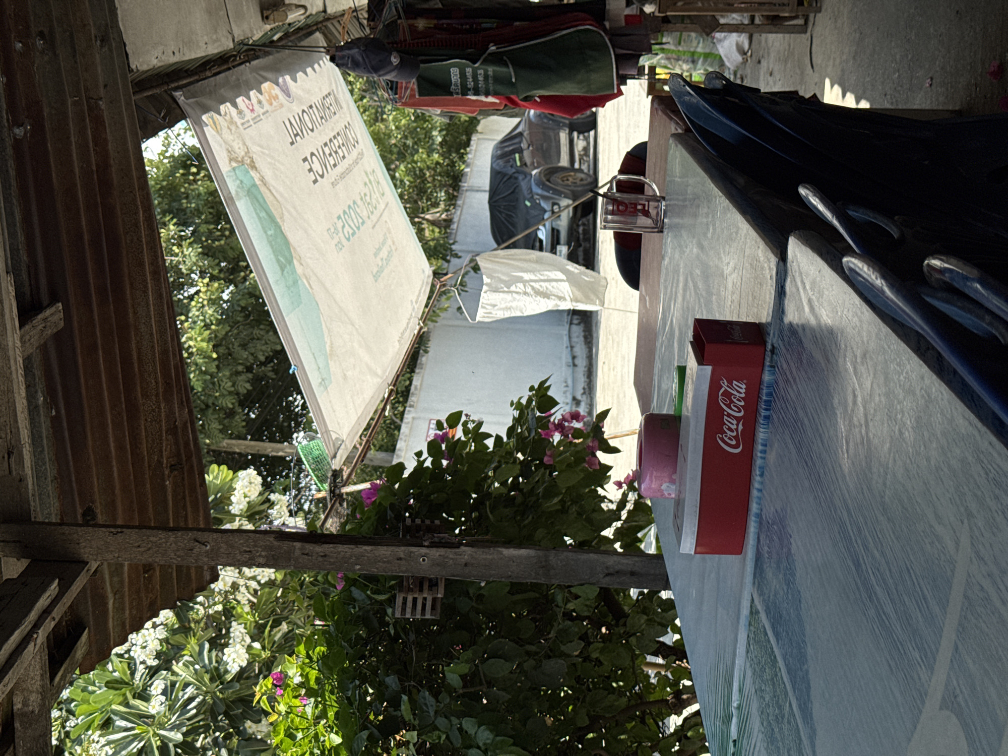 This image captures a vibrant, sunlit outdoor scene, likely a casual eatery or market stall in Pattaya City, Thailand. The setting features a mix of rustic and modern elements, with lush tropical foliage providing a natural backdrop.
In the foreground to the right, a textured blue tabletop, possibly covered with plastic, extends across the frame. A red Coca-Cola box rests on the table, and several blue plastic hangers or hooks are arranged nearby. Further back on the table, a clear glass mug displaying the red "LEO" logo is visible, suggesting a popular Thai beer brand.
To the mid-left, a large white banner is hanging, showing mirrored text that, when read correctly, spells "INTERNATIONAL CONFERENCE" along with dates "16-18 / 2023" and other unreadable Thai or English text, and several small logos. Adjacent to the banner, weathered, corrugated metal sheeting forms part of a structure.
The background is dominated by dense green foliage, including trees and flowering bushes with white and pink blooms, characteristic of Plumeria and Bougainvillea. A car covered by a dark tarp is parked further back, and a white building wall is also visible. The bright lighting and distinct shadows indicate a sunny daytime. The overall atmosphere appears informal and relaxed, hinting at a local gathering or refreshment point.