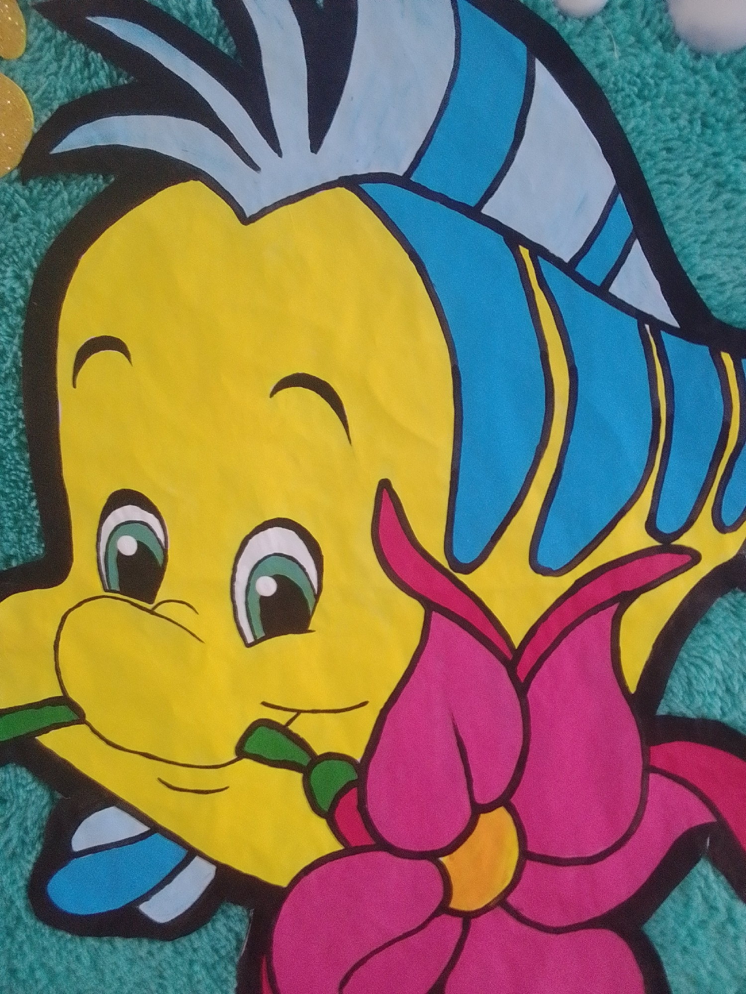 The media file is a close-up, high-angle shot of a colorful, cartoon-style drawing of a yellow fish, possibly Flounder from Disney's "The Little Mermaid." The fish has large, expressive green and white eyes, a simple smiling mouth with a green stem held in it, and blue fins and hair. It is positioned on a teal, fuzzy background, which could be a towel or fabric.
To the right of the fish's head, a large, stylized pink flower with a yellow center is depicted, with its petals overlapping the fish's face and body. The drawing features bold black outlines separating the colors. In the top left corner, a partial yellow circle with gold speckles is visible, suggesting another decorative element in the composition.
The artwork appears to be a craft or decorative piece, possibly a cutout for a child's room or a party decoration. The overall mood is cheerful and playful due to the bright colors and cartoonish style. No text is visible in the image. The location context provided is La Tagua, Venezuela.