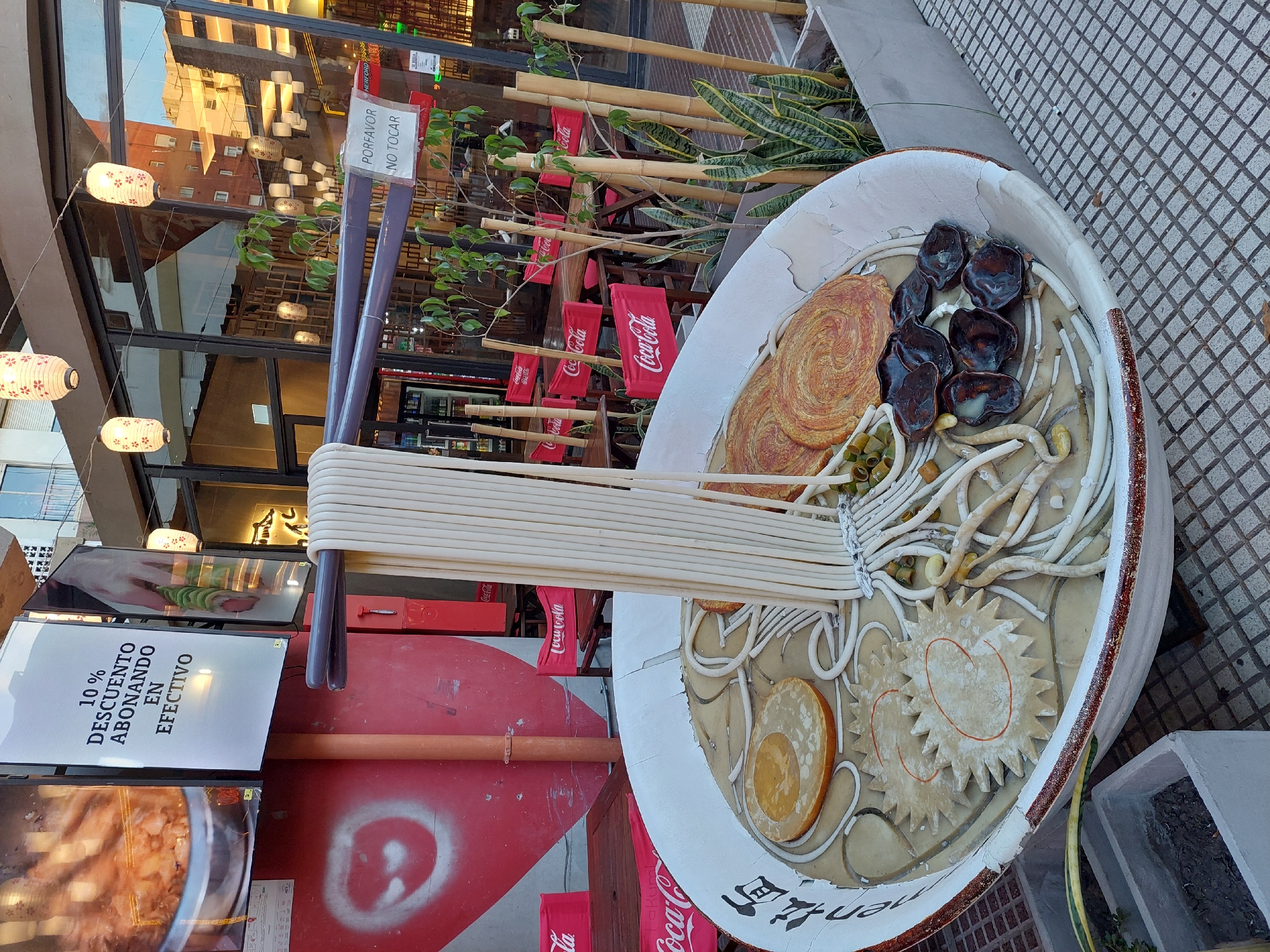 The image displays a large, outdoor ramen bowl sculpture with oversized chopsticks, likely serving as an eye-catching advertisement for an Asian restaurant in Buenos Aires, Argentina. The colossal bowl is filled with highly detailed faux ramen ingredients, including long white noodles being lifted by the chopsticks, dark brown mushroom-like pieces, golden spiral-cut elements, pale yellow rounds resembling eggs or fish cakes, and various small green and light-colored vegetables.
Behind the sculpture is the glass facade of a building, reflecting nearby structures and interior lighting fixtures. A string of lit, traditional Japanese-style paper lanterns hangs above the display. A decorative barrier made of bamboo poles and potted snake plants runs alongside the building, adorned with numerous small red flags bearing the "Coca-Cola" logo.
Visible text includes a digital sign prominently displaying "10% DESCUENTO ABONANDO EN EFECTIVO" (10% DISCOUNT PAYING IN CASH). A small white sign attached to the chopsticks reads "POR FAVOR NO TOCAR" (PLEASE DO NOT TOUCH). The word "Coca-Cola" appears multiple times on the red flags, and "HERFORD" is visible in small text near the top right. The ground is paved with grey, patterned tiles. No people are visible, and the scene presents a static, decorative outdoor display, likely set during the day given the ambient light, though the illuminated lanterns could suggest evening or purely decorative lighting.