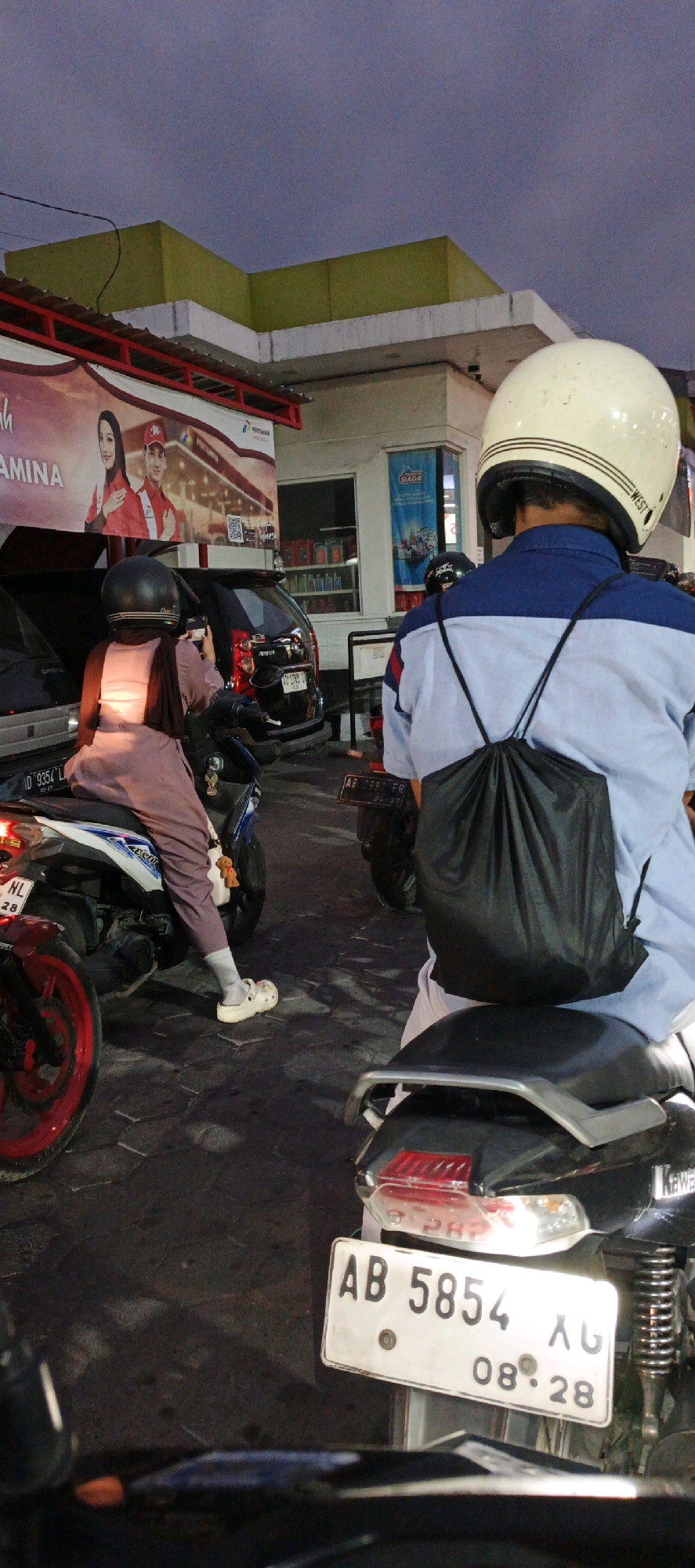 The image captures a scene at a Pertamina gas station in Kasihan, Indonesia. The foreground features the back of a motorcycle with its license plate clearly visible, reading "AB 5854 AG 08-28". To the left, a person wearing a helmet and a light purple outfit sits on another motorcycle, holding a smartphone. Behind them, a black car is parked. In the background, the gas station's canopy and building are visible, along with various products displayed inside. The sky is overcast, suggesting it might be dusk or early evening. Several other motorcycles and individuals are also present, indicating a busy environment at the gas station.