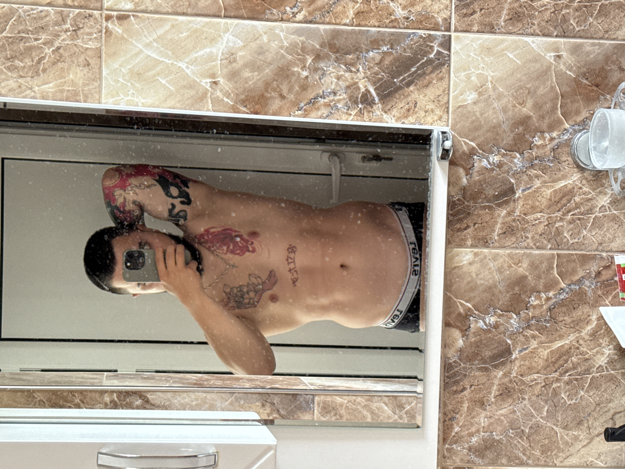 The image displays a mirror selfie taken by a person in a bathroom, with the content rotated 90 degrees counter-clockwise. A shirtless individual with dark hair is seen reflected in a rectangular mirror, holding a smartphone to capture the image. The person's left arm is bent, and their gaze is directed towards the phone.
Numerous tattoos are visible on the person's body: a prominent red and black floral design covers parts of the left shoulder and chest, while what appears to be East Asian characters are tattooed on the upper torso. The person is wearing dark underwear, with the word "REAL" distinctly visible in reverse, in white lettering, on the waistband.
The mirror surface is speckled with what appears to be water droplets or dust. The background outside the mirror consists of bathroom walls clad in brown and beige marble-patterned tiles. To the right of the mirror, a clear plastic toothbrush holder is mounted on the tiled wall. A portion of a white vanity unit with a silver handle is visible below the mirror. The setting is an indoor bathroom. The location context provided for this image is Plovdiv, Bulgaria.